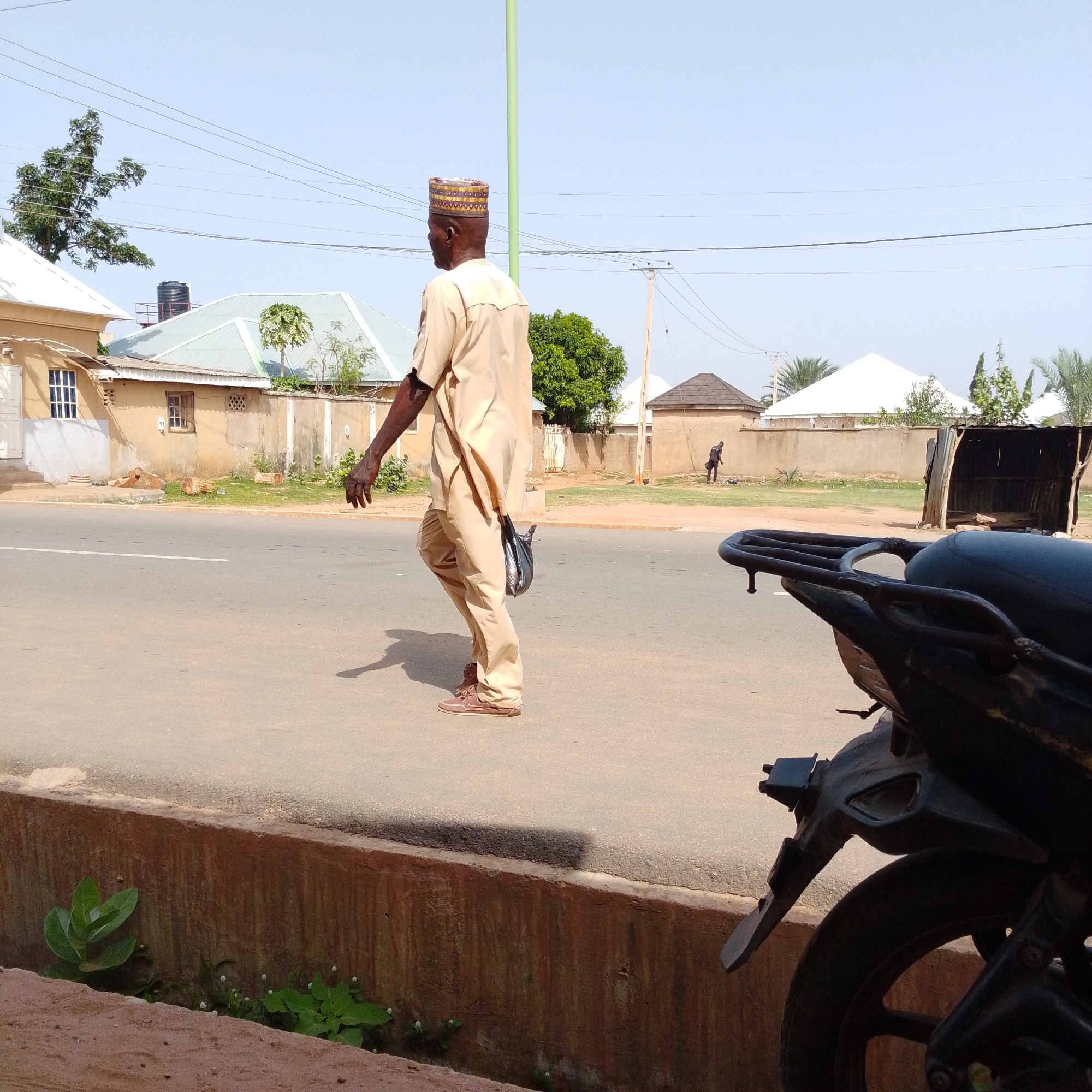 In Gombe, Nigeria, a man wearing a light beige traditional outfit and a patterned cap walks on the right side of a paved road, facing away from the camera. He is carrying a black plastic bag in his right hand. The road has a white line marking its center. To the left of the road is a raised concrete curb with some green vegetation growing in front of it. Behind the man, a few buildings with light-colored walls and pitched roofs are visible, along with trees and overhead power lines against a clear blue sky. In the foreground on the right, the back of a black motorcycle is visible. In the background, another person is seen near a structure.