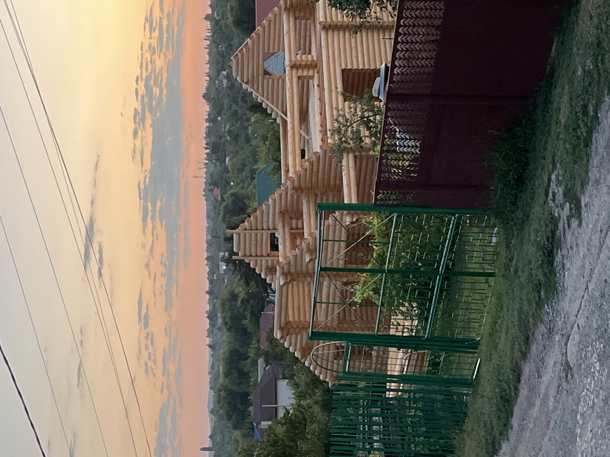 This image depicts a construction site of a large wooden house, likely a log cabin, at sunset in Saratov, Russia. The house is in the process of being built, with exposed logs forming its walls and roof structure. A green metal fence runs across the foreground, partially obscuring the view of the construction. Behind the fence, a dark red fence runs parallel to it. The sky displays hues of orange and pink, characteristic of a sunset, with scattered clouds. Power lines are visible against the sky in the upper portion of the image. In the background, a dense green landscape with trees and other buildings suggests a rural or suburban setting. There are no people or animals visible in the image. The overall scene is peaceful and evokes the end of a day's work.