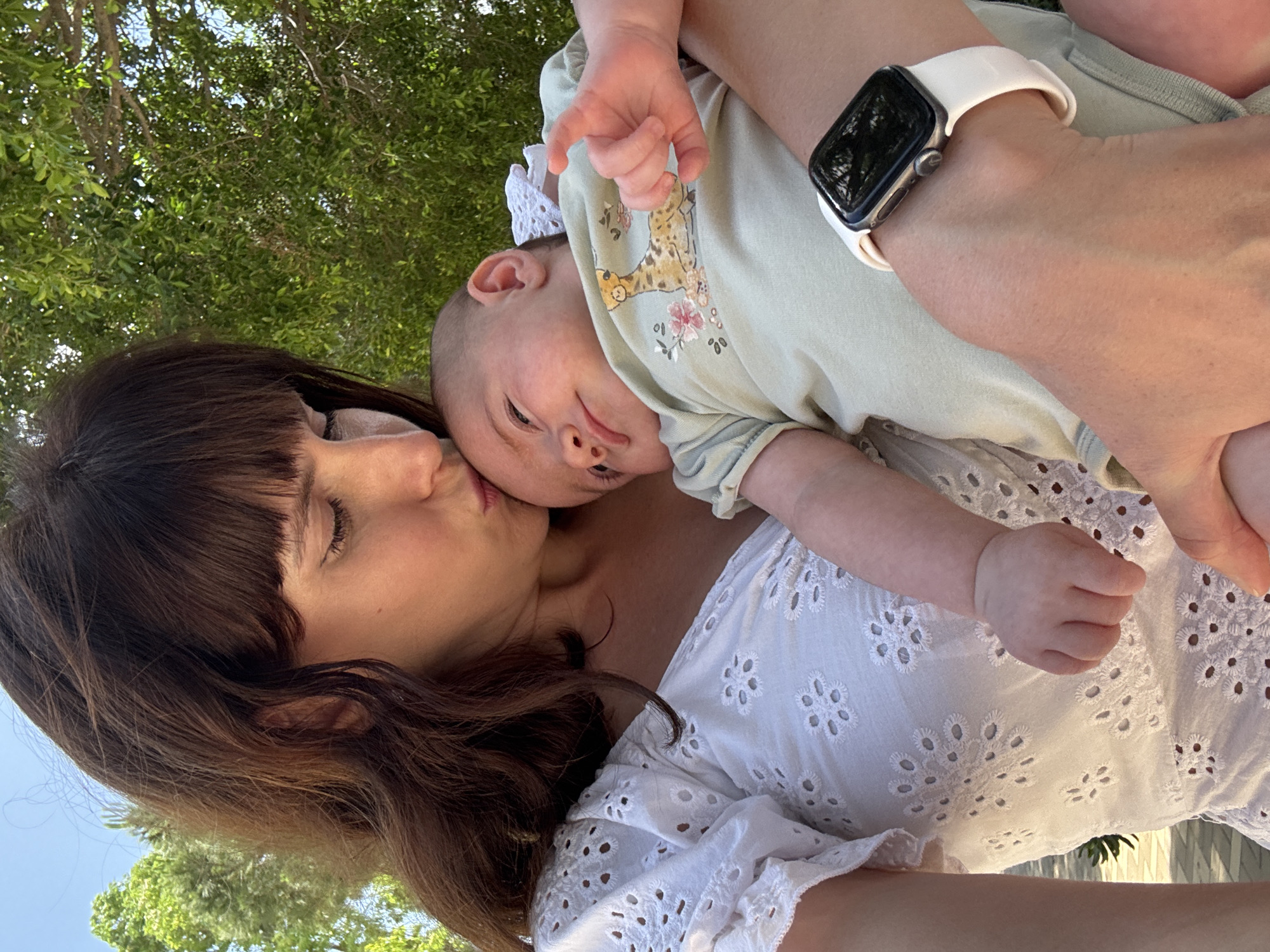 A close-up, medium shot shows a woman in a white eyelet top kissing a baby's forehead. The woman has dark brown hair with bangs and is wearing a white top with eyelet detailing. The baby is wearing a light green onesie with a giraffe print and floral accents. The baby's right hand is reaching up towards the woman's face. A white Apple Watch is visible on the woman's left wrist. The background consists of green trees and a bright blue sky, suggesting an outdoor setting. The location is identified as Isola delle Femmine, Italy.