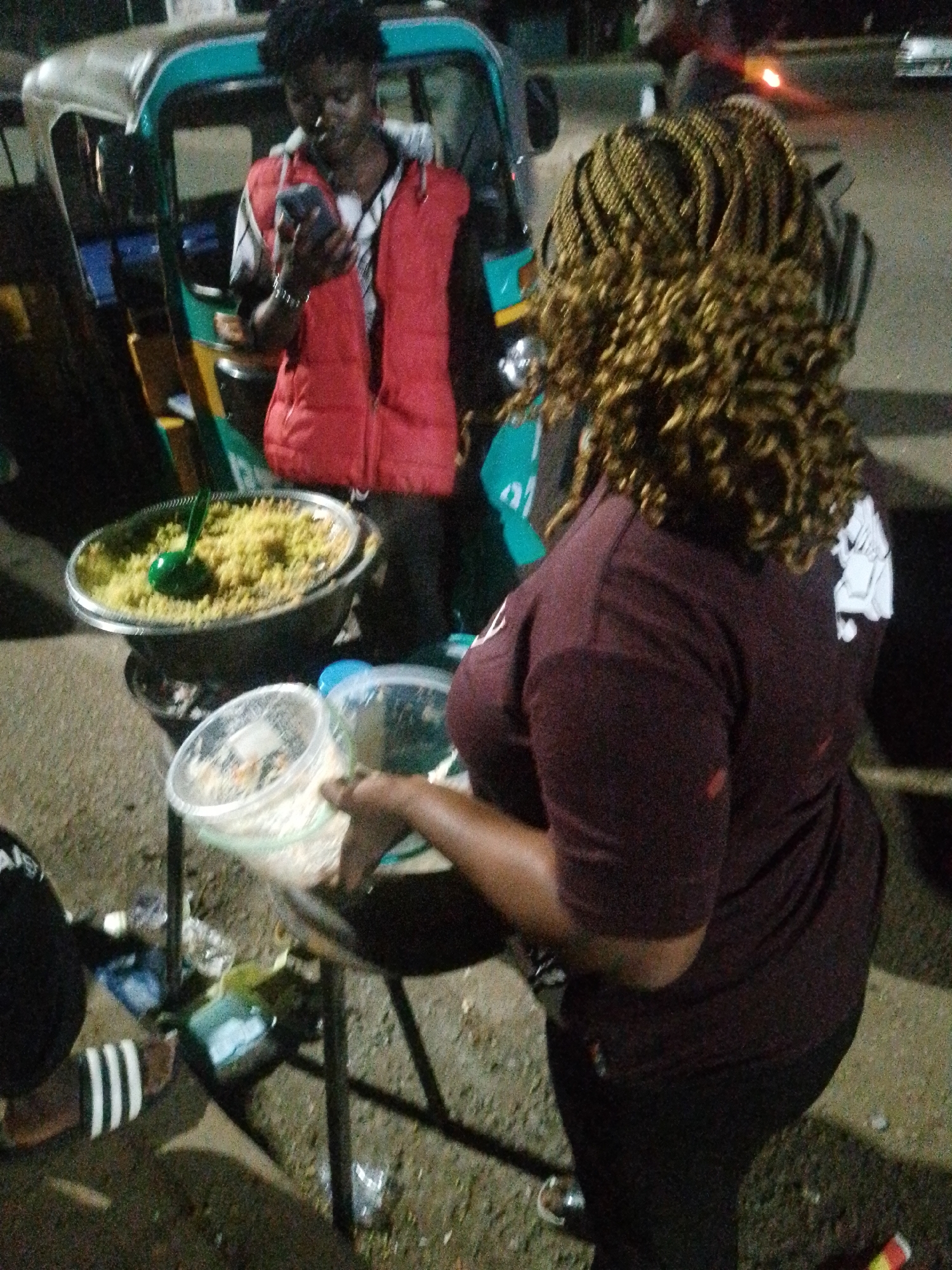 In an outdoor, nighttime urban scene, a person wearing a red vest over a checkered shirt stands by a three-wheeled vehicle, looking at a mobile phone. Beside them, a large metal bowl filled with yellow rice sits on a stand. A green serving spoon is placed in the bowl. In the foreground, another person, viewed from the back, has elaborate blonde braids and wears a maroon t-shirt. They are holding a clear plastic container filled with a lighter-colored food item, possibly rice or couscous. The ground is gritty and appears to be dirt or gravel, with scattered plastic bottles and debris. The lighting is dim, suggesting artificial light sources are present.