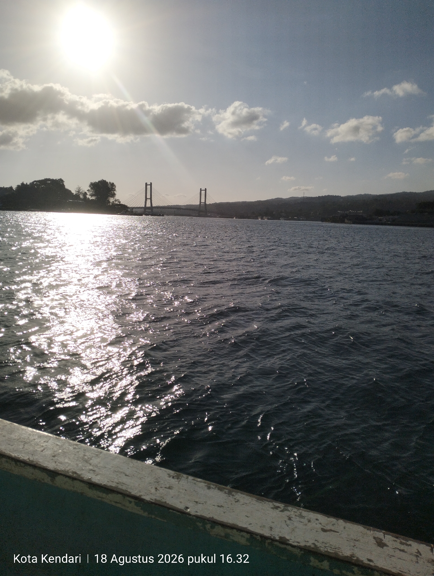 The image shows a body of water, likely a bay or strait, with a distant bridge. The sun is bright and positioned in the upper left quadrant, casting a strong glare on the water's surface. Clouds are present in the sky. The left side of the image shows a body of water with significant glare from the sun. In the distance, there is a silhouette of land with trees, and a suspension bridge with two towers. The right side of the image shows the open water with a dark, rippling surface. In the foreground, the edge of a vessel is visible, with a weathered wooden railing. Text overlays the bottom of the image: "Kota Kendari | 18 Agustus 2026 pukul 16.32", indicating the location as Kendari City, and the date and time as August 18, 2026, at 4:32 PM.