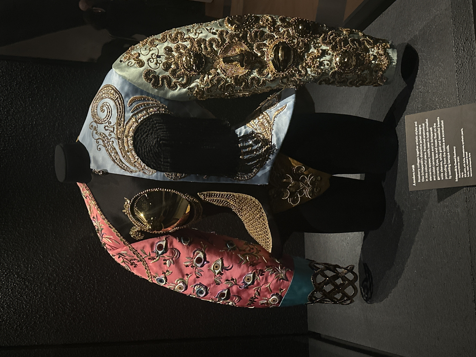 A highly embellished jacket is displayed on a black, headless mannequin within a dark museum exhibition space. The jacket is bisected into two distinct designs: the viewer's left side features a pink fabric sleeve embroidered with multiple human eye motifs and green leafy patterns, terminating in a teal cuff with a metallic, open-weave gold-colored band. The viewer's right side features a light blue-green fabric sleeve intricately embroidered with gold baroque scrolls and embedded three-dimensional elements resembling eyes, lips, and noses. The jacket's bodice includes a large, polished gold oval resembling a mirror on the left and a textured black, possibly tasseled, component on the right. The mannequin stands on a dark grey platform against a dark, textured wall, illuminated by spotlights. A black exhibition plaque at the bottom right identifies the piece as 'Schiaparelli by Daniel Roseberry, Autumn/Winter 2021, Paris,' and details its materials and design influences related to Elsa Schiaparelli. This object is exhibited at the Victoria and Albert Museum (V&A) in London, United Kingdom.