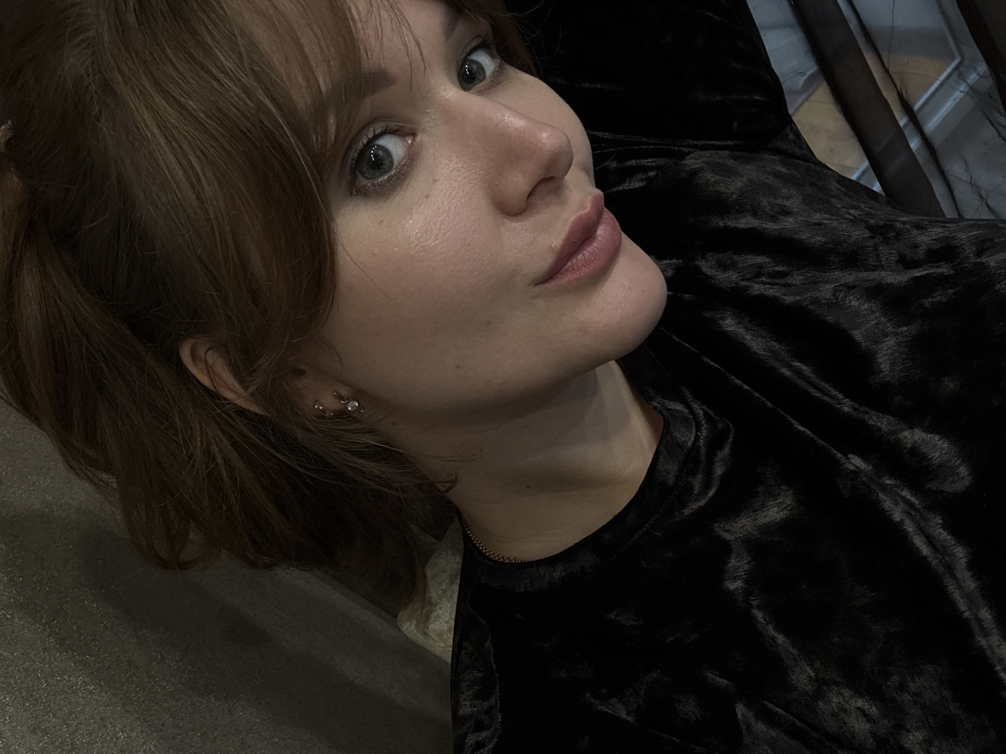 This media file is a close-up, high-angle selfie of a young woman lying down. She has light brown hair pulled back into two pigtails and blue-green eyes. She is wearing a black velvet top and a delicate gold chain necklace. A small hoop earring with a clear stone is visible on her left ear. The background is dark and indistinct, but suggests an indoor setting, possibly a home. The lighting is dim, creating soft shadows on her face. The woman has a neutral expression, with her lips slightly parted. The overall mood is relaxed and intimate. There is no visible text in the image.