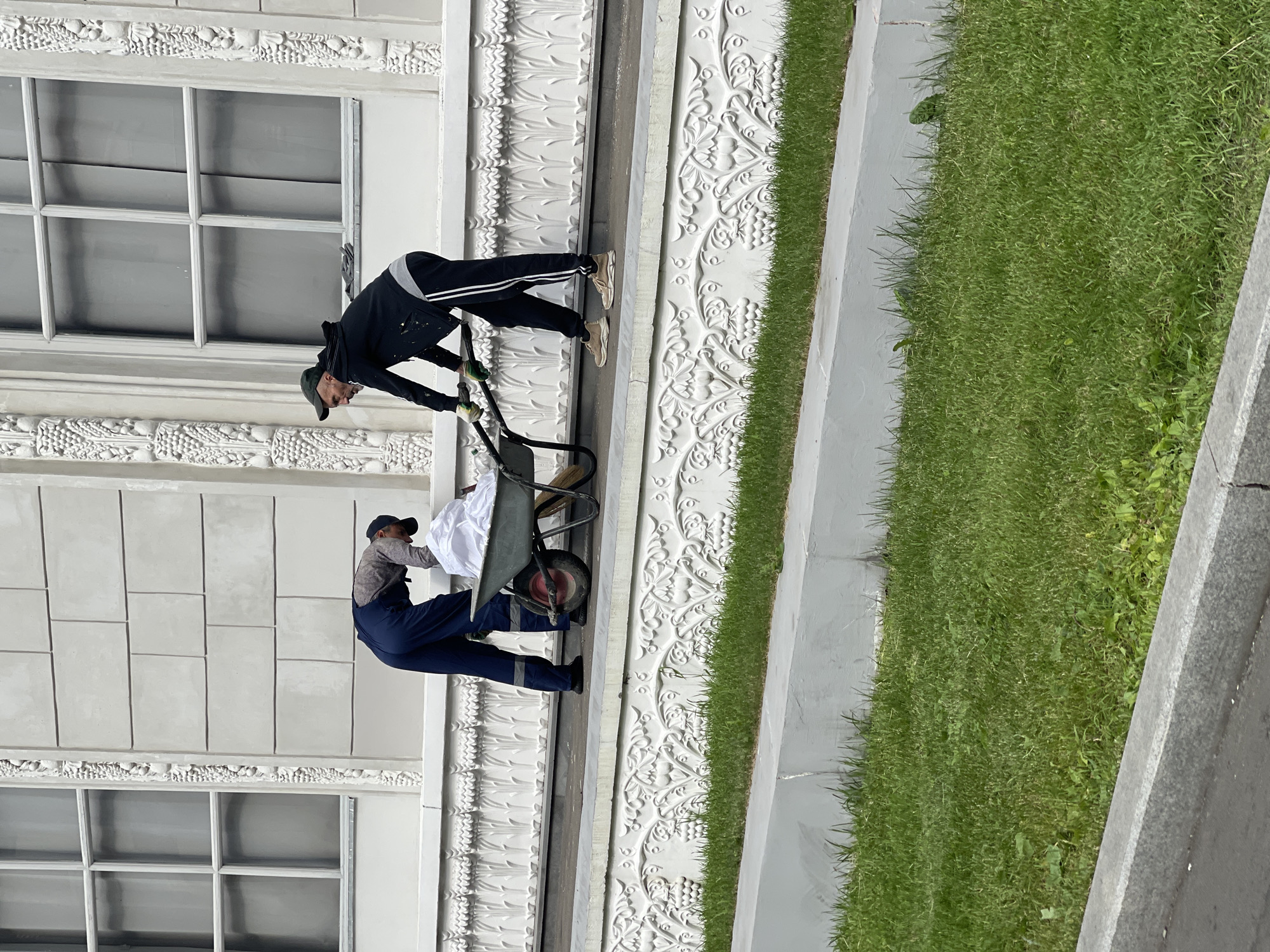 Two individuals are engaged in work on an elevated white platform in front of a large, ornate white building. The building features a row of tall, multi-paned windows and decorative architectural elements resembling grapes and leaves. One person, wearing blue overalls with reflective stripes, is bent over a wheelbarrow, appearing to be loading or arranging white material within it. The second person, dressed in dark athletic wear and a baseball cap, is pushing the handles of the wheelbarrow, suggesting they are assisting in moving it. The wheelbarrow contains a significant amount of white material, possibly fabric or cleaning supplies, and a broom is visible resting against its side. The platform is bordered by a white, decorative ledge with a floral motif, and a strip of green grass is situated below this ledge. Further down, a concrete border runs parallel to a paved surface. The scene appears to be outdoors in daylight.