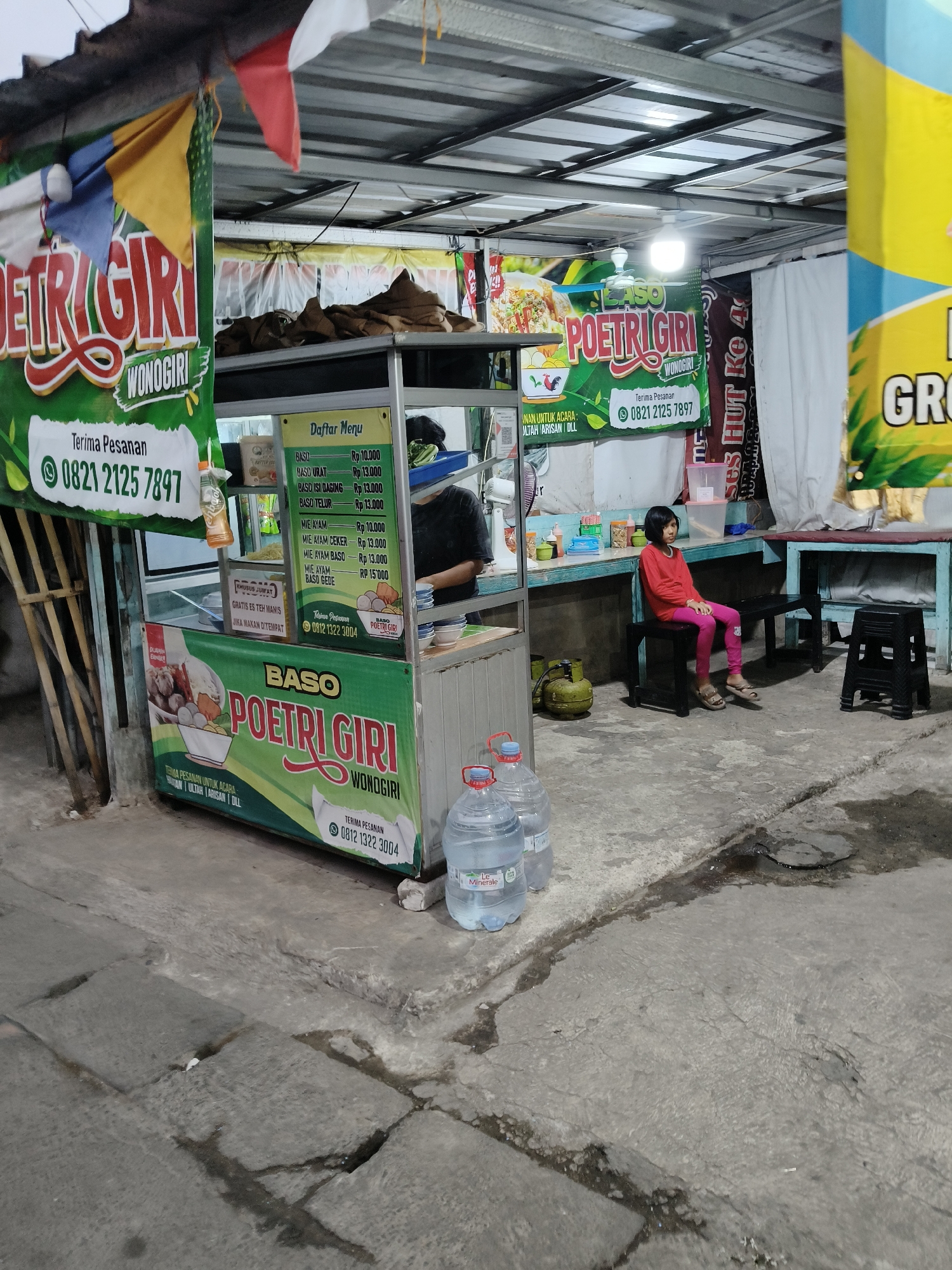 The image depicts an outdoor food stall operating at night. The stall, identified by banners with the text "BASO POETRI GIRI WONOGIRI", appears to be selling meatballs ("baso"). A menu board is visible, listing various meatball dishes and prices in Indonesian Rupiah. A young girl, wearing a red shirt and pink pants, sits on a dark bench to the right of the stall. Two large bottles of drinking water are placed on the ground in front of the stall. The stall is situated on a concrete pavement, with some areas appearing wet. A propane gas cylinder is visible next to the stall. The background includes a corrugated metal roof, white fabric curtains, and another food stall banner with yellow and blue colors. The overall scene suggests a bustling, albeit simple, commercial setting common in urban areas of Indonesia.