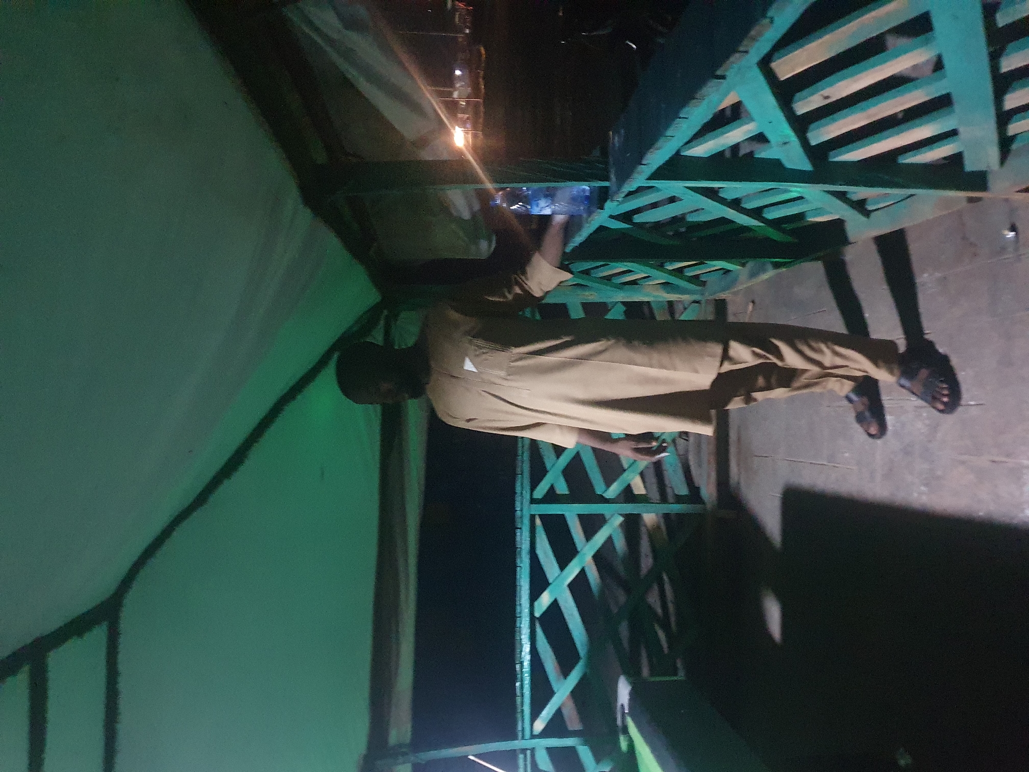 A full-body shot captures a man standing on a wooden deck under a green awning. He is wearing a tan, knee-length traditional outfit and dark sandals. His right hand rests on a light blue plastic bottle of water, placed on the green railing. The railing is made of crisscrossed wooden planks. The man is facing slightly to his right, looking out at the dark, blurry background which appears to be an outdoor setting at night, with some distant lights. The awning overhead is a translucent green fabric supported by dark beams. The ground beneath is paved with concrete or stone tiles. The overall ambiance suggests a rest stop or observation point, possibly in a park or near a waterfront. The scene's lighting is dim, with the awning casting a green hue. This scene is located at the Lokoja Confluence Park in Lokoja, Nigeria, a location known for the confluence of the Niger and Benue rivers.