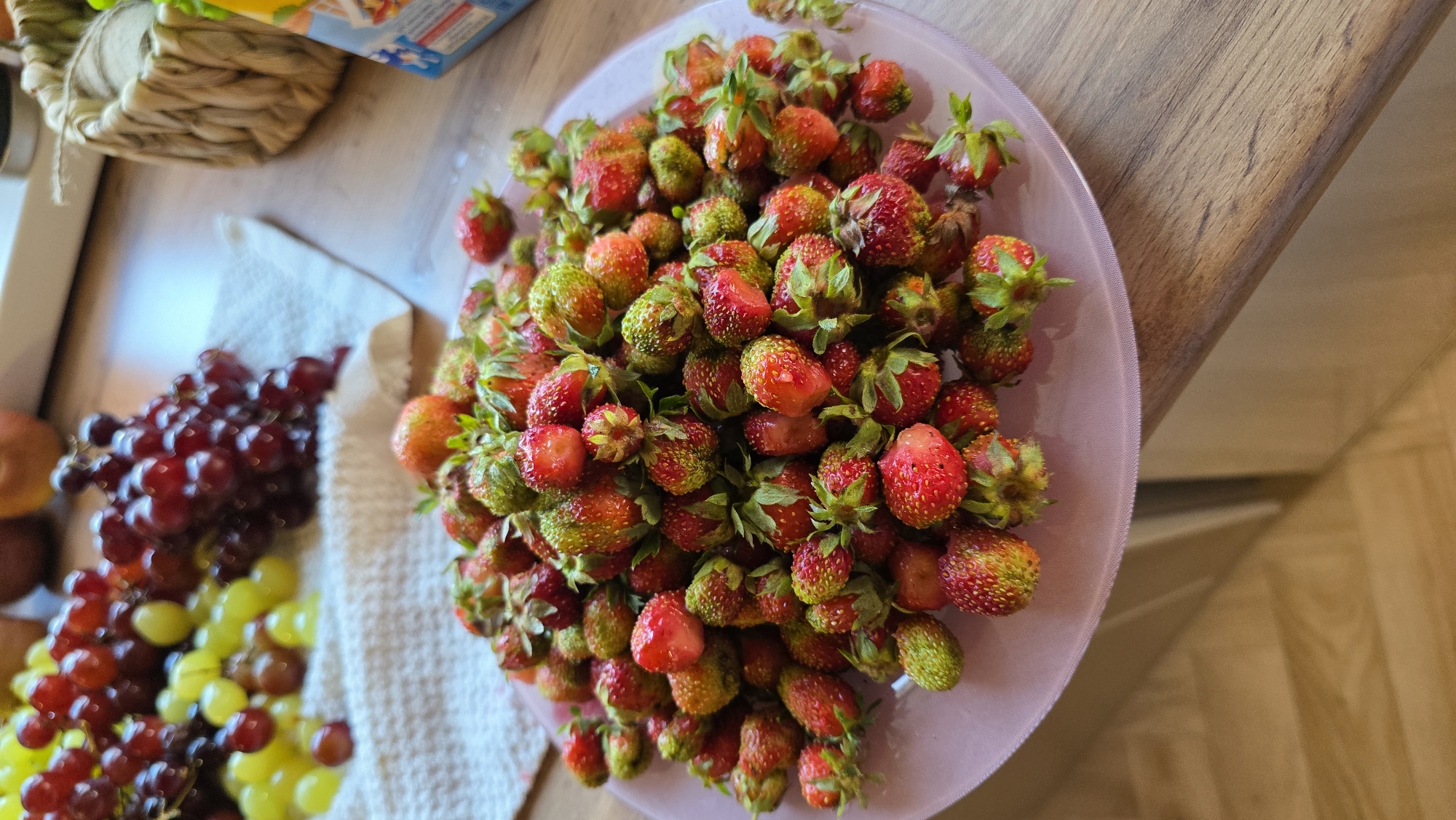 This image captures a close-up overhead view of a pile of fresh strawberries, predominantly red with some green hues, resting on a light pink plate. The strawberries are small and appear to be wild or miniature varieties, with visible green stems and seeds. Some strawberries show cuts, revealing their juicy interiors.
To the left of the plate of strawberries, a cluster of red grapes and a bunch of green grapes are visible, also appearing fresh. These grapes are on a white cloth or napkin. Further to the left, a woven basket with twine wrapping is partially visible, suggesting a rustic or natural setting. In the background, a wooden surface, likely a table or countertop, is visible, with a hint of flooring. A portion of a colorful box, possibly containing food items, is also in the upper left corner of the frame.
The lighting suggests daytime, possibly indoors, with soft shadows. The overall scene evokes a sense of freshness and abundance, likely a home setting where fruit has been recently gathered or prepared. There is no visible text in the image that can be clearly read. The crops provide closer views of the strawberries and grapes, highlighting their textures and colors, and also give a better view of the surrounding objects and surfaces.