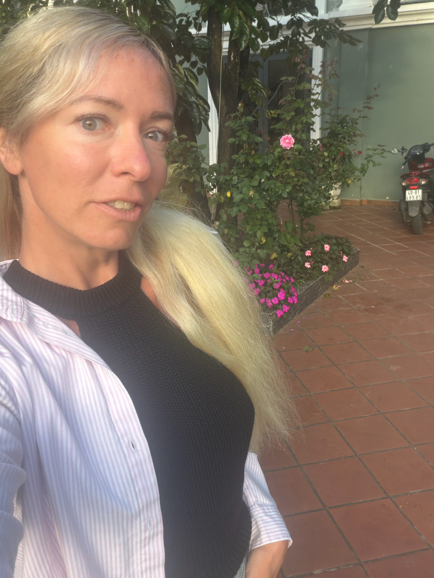 A blonde woman is taking a selfie outdoors. She is wearing a pink and white striped button-down shirt over a black knit top. Behind her, there is a rose bush with a pink rose in bloom, a tree with green leaves, and a wall painted light green. A red scooter is parked to the right of the frame, with a license plate that reads "49-B1 489 85". The ground is paved with reddish-brown square tiles. The scene is lit by natural light, casting soft shadows.
