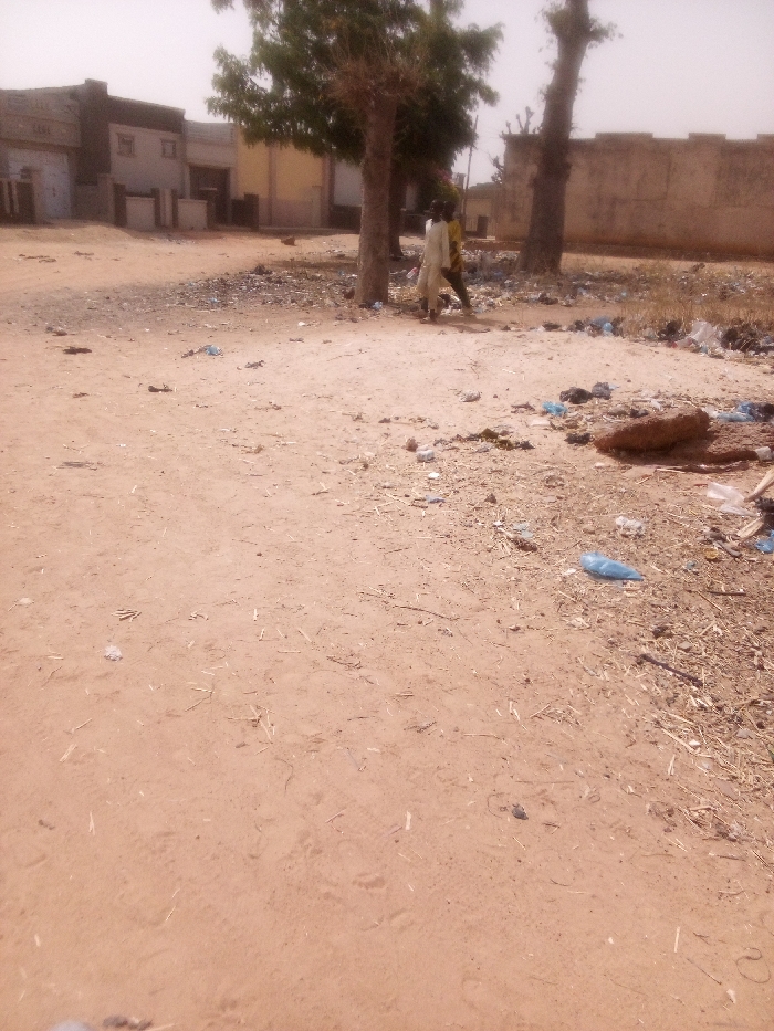 The image depicts an outdoor scene in Tudun Murtala, Nigeria, characterized by a predominantly reddish-brown, sandy dirt ground covering the foreground and mid-ground. The ground is uneven and littered with various pieces of refuse, including blue and white plastic bags, wrappers, and dried organic debris. In the mid-ground, two individuals are present near two prominent trees with visible trunks and sparse green and dry foliage. One individual wears a long, light-colored, possibly white or cream, garment and stands slightly ahead of the second individual. The second individual wears a darker, possibly yellow or ochre, garment. Both individuals appear to be walking or standing, facing generally towards the right side of the frame. In the background, multiple single and multi-story buildings with various architectural styles are visible on the left, alongside a tall, light-colored wall extending across the right. The sky is hazy and bright, indicating daylight conditions.