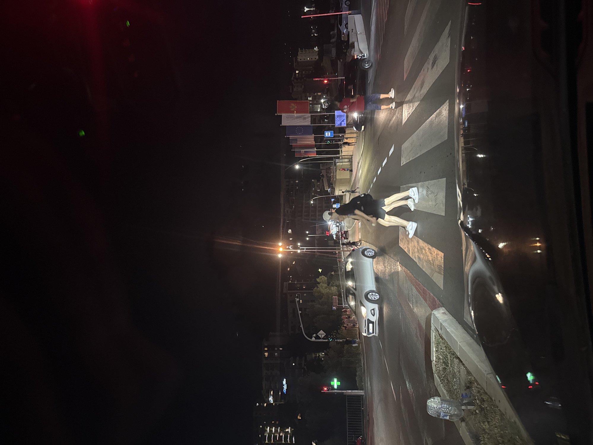 The image captures a night scene on a street in Boreti, Montenegro, focusing on a pedestrian crossing. Two individuals, a male and a female, are walking across the crosswalk from left to right. The male is wearing a white cap, dark shorts, and white sneakers. The female is wearing a black backpack, dark shorts, and white sneakers. A white car is stopped at the crossing, yielding to the pedestrians. In the background, several flags are displayed, including the Montenegrin flag, the US flag, and what appears to be the flag of the European Union. Buildings with illuminated windows are visible in the distance, suggesting an urban environment. Streetlights and traffic signals illuminate the road, casting reflections on the wet surface of the street. To the far right, another individual in a red shirt and blue shorts is standing on the sidewalk, and a white car is parked. The foreground shows the interior of a vehicle looking out at the scene.