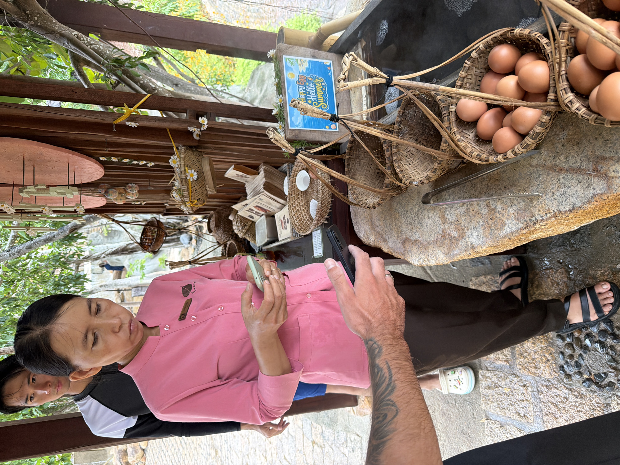 A woman wearing a pink uniform and dark pants is holding two mobile phones, looking down at them. A man with tattoos on his arm is also holding a mobile phone. Behind the woman, another man in a black and white shirt is visible. They are standing outdoors near a display of woven baskets filled with eggs, as well as other decorative items. A sign that says "Happy Egg Hello Summer" is also displayed. The ground is made of stone and gravel, and there are trees and foliage in the background. The setting appears to be a marketplace or a tourist attraction in Nha Trang, Vietnam.