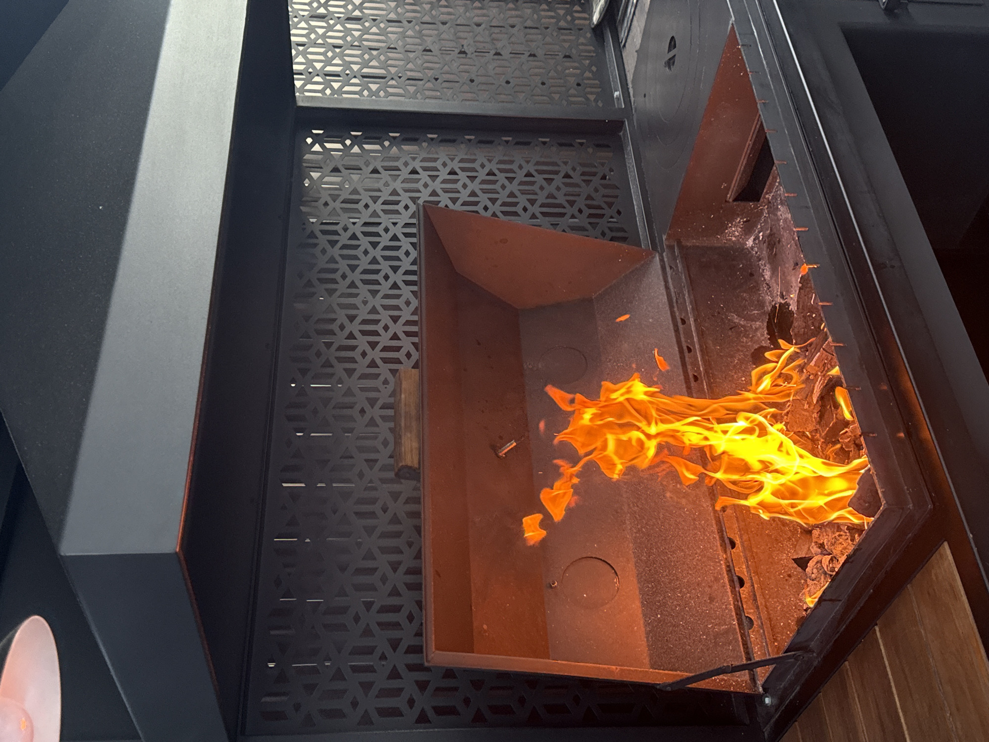 The image shows a close-up view of a large, modern outdoor grill with a fire burning inside. The grill is made of dark metal and has a hinged lid that is open, revealing a fire pit filled with glowing embers and flames. The background features a decorative metal screen with a geometric pattern. The overall scene suggests an outdoor cooking area, possibly a patio or backyard. The location is identified as Starokorsunskaya, Russia.