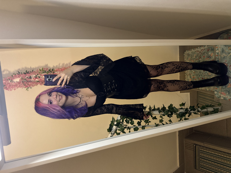 The image displays a full-body mirror selfie taken indoors. A young female individual with wavy purple and pink hair is depicted, smiling at the camera. She is wearing a black lace-sleeved dress or top with a sweetheart neckline, a black corset-style belt, black lace-patterned tights, and black platform boots. Multiple necklaces are visible, and she is holding a dark-colored smartphone, consistent with an iPhone 14 Pro/Pro Max, in her right hand to capture the reflection.
The background consists of a plain, light yellow wall. Artificial green ivy is draped around a white vertical element on the left side of the mirror's reflection, and artificial pink flowers hang vertically on the wall behind the individual's right shoulder. A white rectangular light fixture is mounted above the mirror. The floor beneath the individual's feet is covered by a patterned rug with a light green/teal background. To the left, a striped rug is visible on a lower step or landing, suggesting an indoor stairwell or multi-level area.
No identifiable external landmarks or unique architectural features are present to determine a specific location within the City of Zagreb, Croatia. The setting appears to be a private interior space.