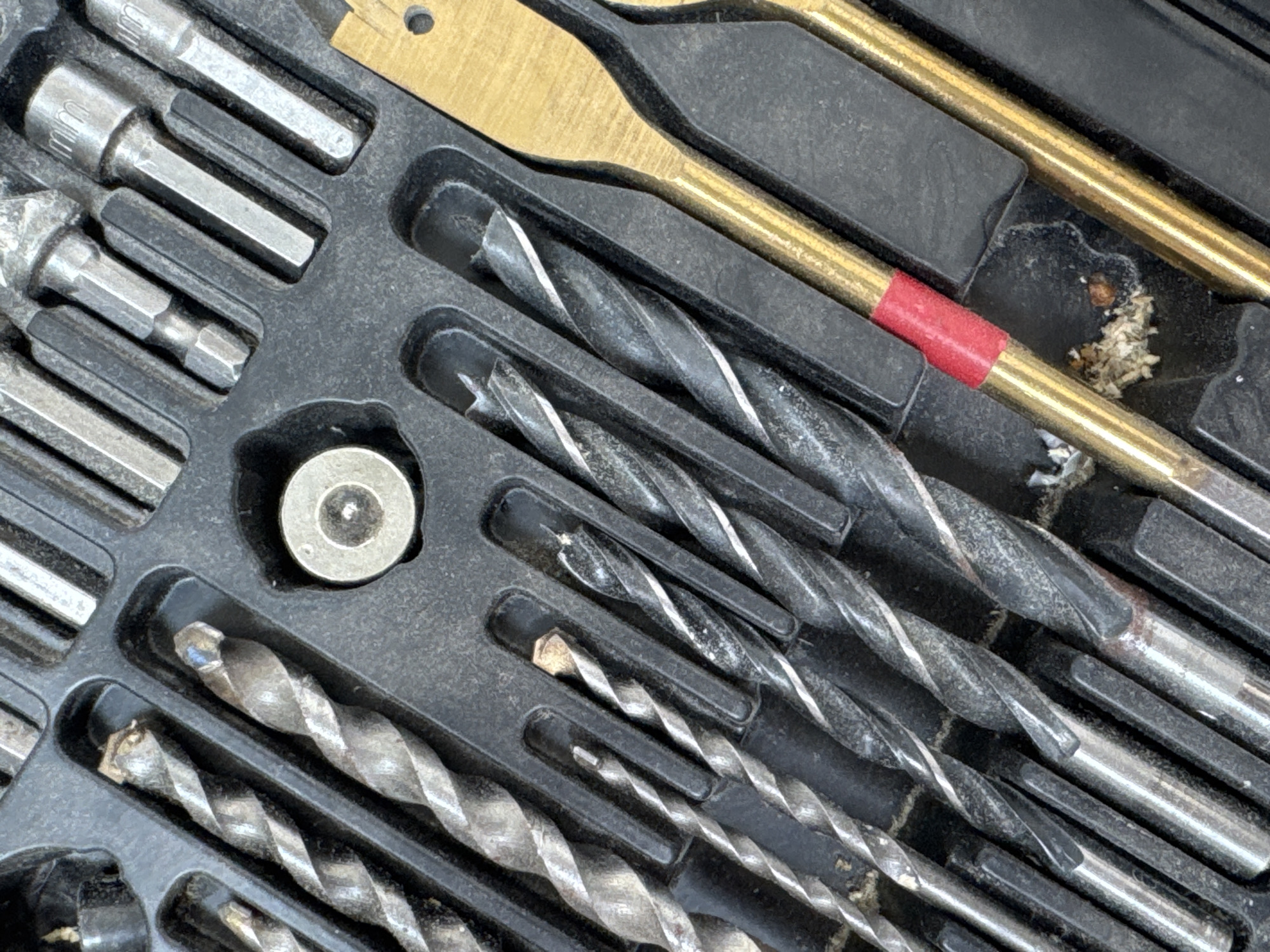 The image is a close-up, top-down shot of a black plastic case filled with various drill bits. The drill bits are made of metal and have a spiral shape, with some appearing to be made of steel and others of a brass-like material. Some of the drill bits are of different sizes and have distinct tips. There are also some hex socket bits in the case. The case itself appears to be worn and slightly dirty, with some debris visible. In the bottom right of the image, there are two gold-colored drill bits, one with a red band around it. The overall impression is that this is a tool organizer that has been used. The image does not contain any information that would allow for the determination of its location in the city of Яромінскі сельскі Савет, Belarus.