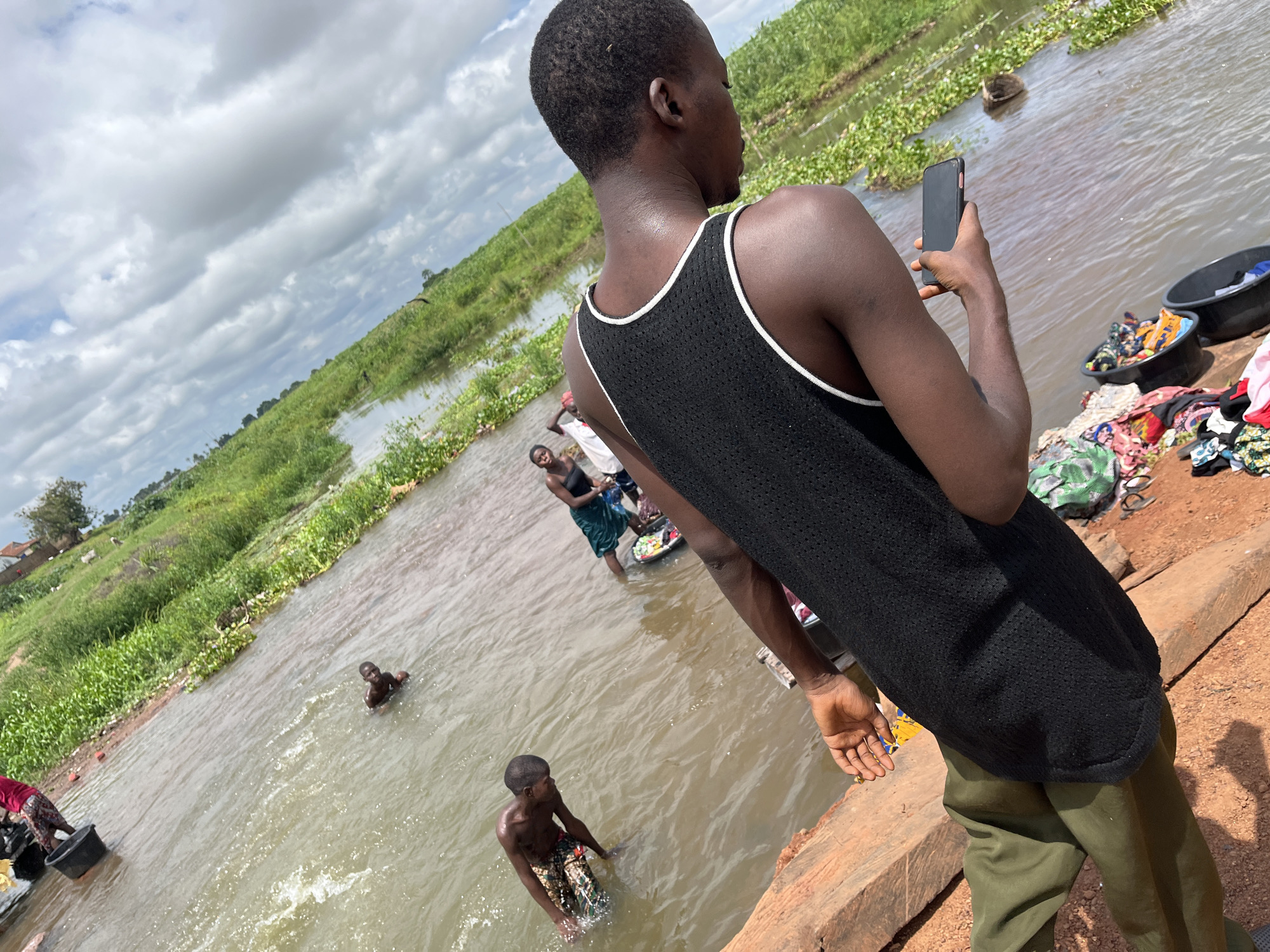 The image depicts a scene in Idah, Nigeria, centered around a body of water, likely a river or stream, under a cloudy daytime sky. In the foreground, a person is seen from behind and slightly to the right, wearing a black mesh tank top with white trim and olive green pants. This individual holds a black smartphone in their right hand, looking out towards the water.
To the right of this person, on the reddish-brown earth bank, are piles of colorful clothing and two dark wash basins, one containing clothes. The water itself is rippled and lined with dense green vegetation, both on the banks and floating on the surface in some areas.
Several other individuals are present in or near the water. Further into the water, a person wearing a patterned garment is partially submerged, appearing to be washing or bathing. Further back and to the left, another individual stands in the water, seemingly engaged in laundry with a basin. A third, smaller figure is seen partially submerged further away in the water. The background consists of lush green growth along the far bank and a wide expanse of grey, cloudy sky. No text is visible in the image.