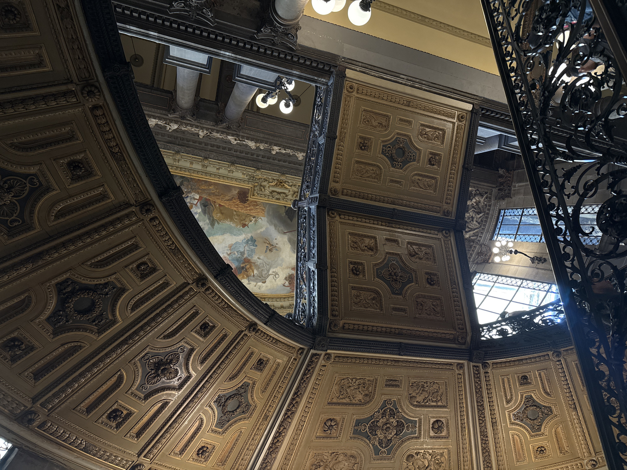 The image shows an interior view of the Palacio de Correos de México (Postal Palace) in Mexico City, Mexico, looking upwards. The architecture is ornate and features a series of elaborately decorated ceilings. Visible is a semi-circular, coffered ceiling with gold and blue detailing. Beyond it is a square, tiered ceiling with similar ornamentation. A mural featuring figures in the sky is partially visible, suggesting a celestial theme. A wrought-iron staircase is present on the right side of the frame, providing a contrasting texture against the decorative ceilings. Light fixtures are visible in the upper portion of the frame, as well as natural light streaming through windows in the distance.