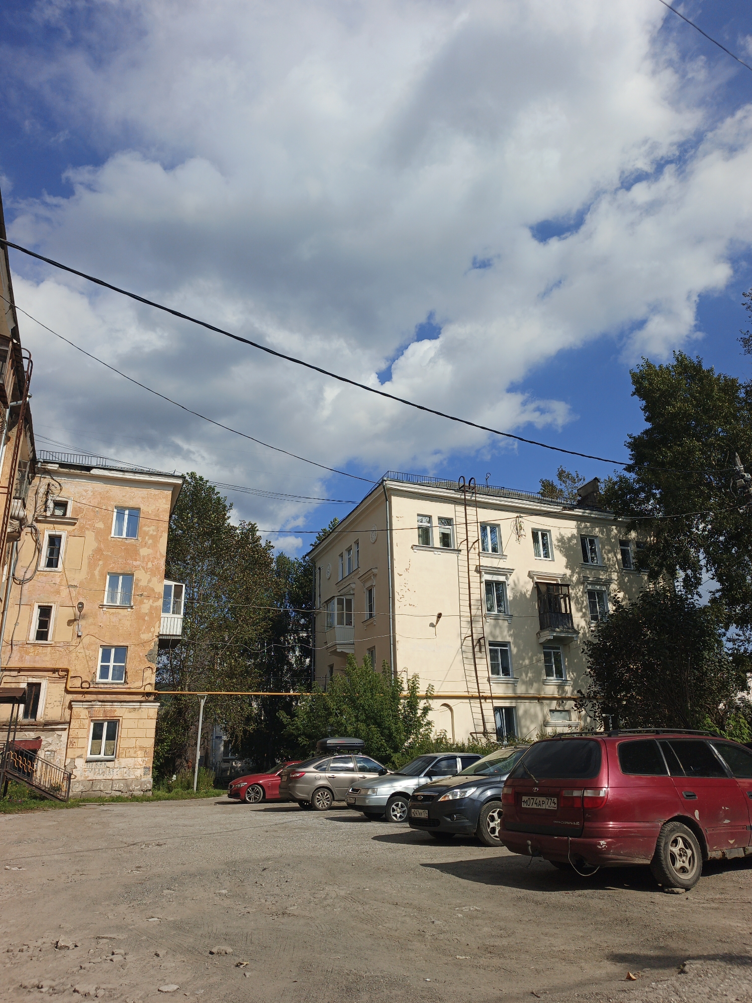 The image depicts an outdoor scene in the city of Zlatoust, Russia, on a sunny day with scattered clouds. The foreground is a dirt and gravel parking lot with several cars parked. In the mid-ground, there are two apartment buildings. The building on the left is older, with a peeling orange facade and visible yellow pipes. The building on the right is a lighter color and appears to be in better condition. Trees are visible between and behind the buildings. Overhead power lines are strung across the sky. The overall impression is of a residential area in a Russian city.