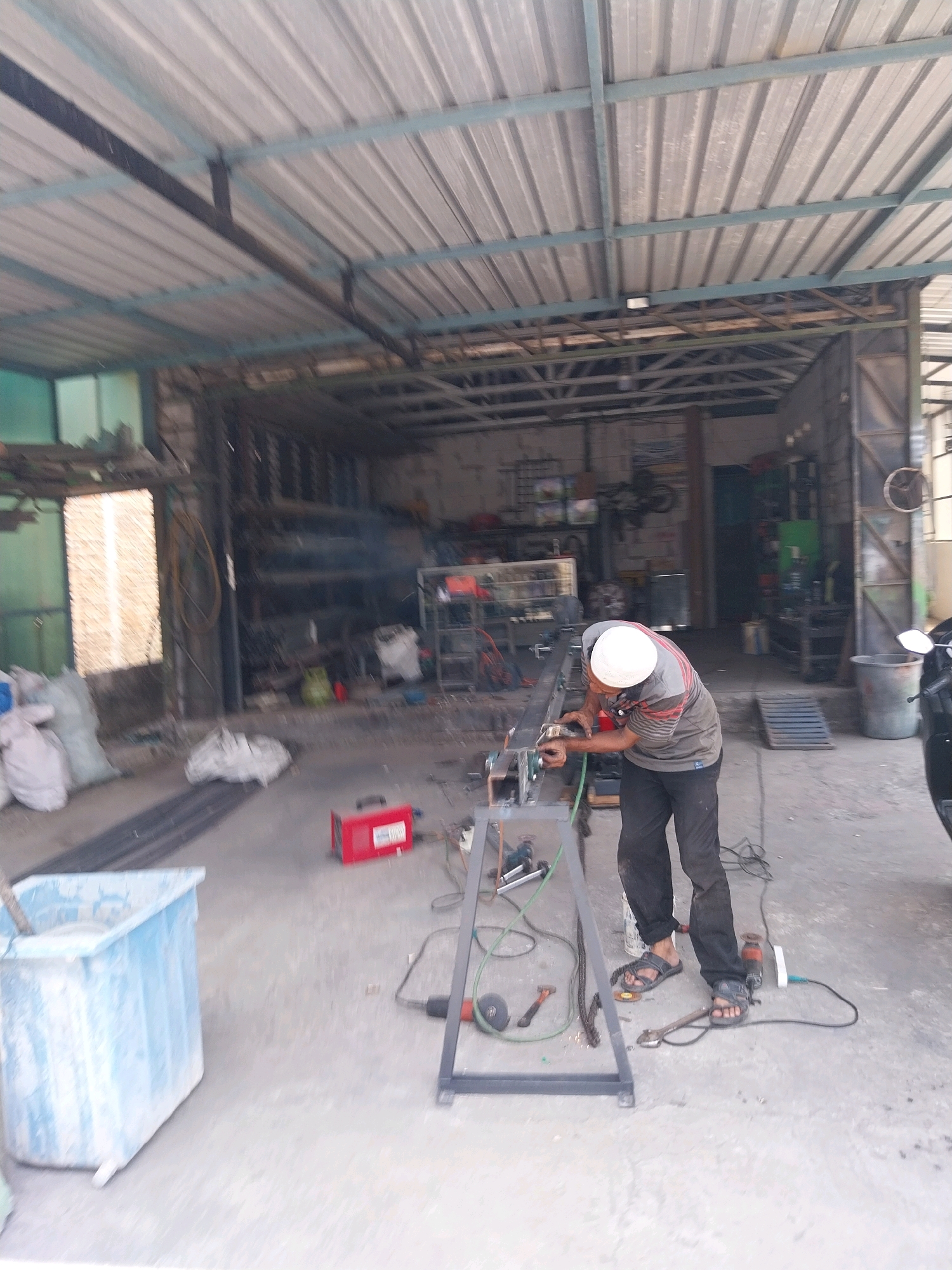 A man is welding metal in a workshop in Campur, Indonesia. The man is wearing a white hard hat and a grey shirt with red stripes. He is bent over a metal frame, working on it with a welding torch. Sparks are flying from the welding torch. There are several other objects in the workshop, including a red welding machine, a blue plastic tub filled with liquid, and bags of material. The workshop appears to be open-air, with a corrugated metal roof supported by metal beams. The floor is concrete. In the background, there are shelves stocked with various items, and a motorcycle is parked to the right of the frame.