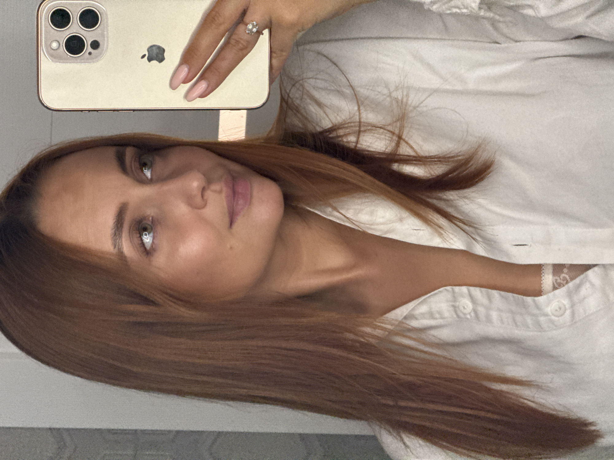 The image is a close-up selfie of a woman with auburn hair, taken in what appears to be a brightly lit indoor space. She is wearing a white button-down shirt, partially unbuttoned to reveal lace trim of an undergarment. Her right hand, adorned with a diamond ring and a pale pink manicure, holds a gold iPhone to take the picture. The background shows a textured wall and a hint of what could be a mirror or reflective surface. There is no indication of the city of Chaikovsky, Russia, in this image.