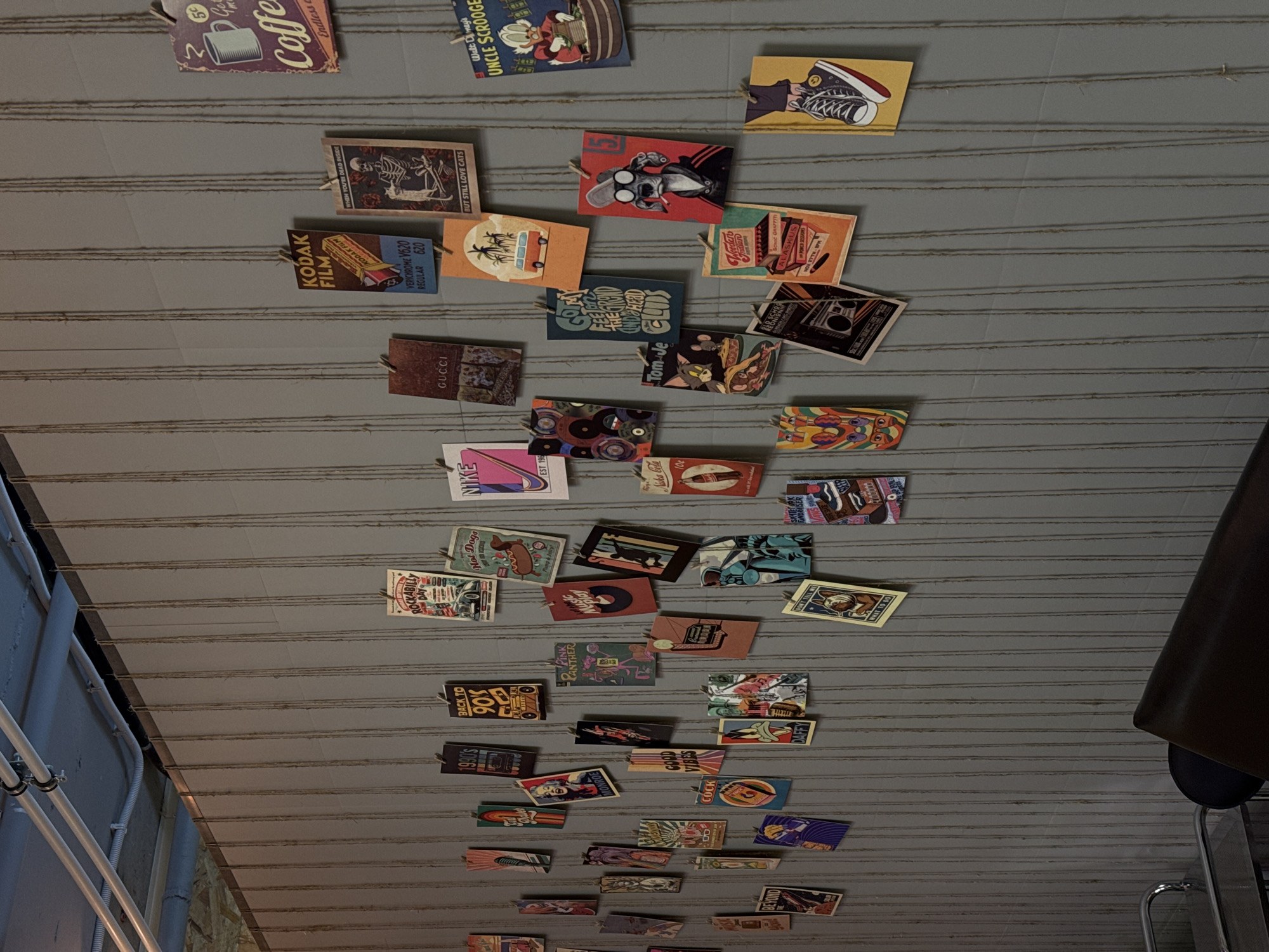 An indoor, overhead shot captures a dense display of small, rectangular, retro-themed cards or tags hanging from strings against a light gray, grooved surface, likely a ceiling or wall. The cards are attached to strings with small clips, creating an eclectic, decorative arrangement.
The subjects on the cards vary widely, encompassing pop culture references, vintage advertisements, and cartoon characters. Visible items include brand logos like "Kodak FILM" (with "VERICHROME V620 REGULAR 620"), "GUCCI," "NIKE," and "Coca-Cola." Cartoon characters such as Uncle Scrooge, Tom and Jerry, Pink Panther, and Daffy Duck are featured. Other themes include "BACK TO 90's" with a boombox, Marilyn Monroe, a skeleton with text "WHEN YOU'RE DEAD INSIDE BUT STILL LOVE CATS," and various vintage-style illustrations like hot dogs, a classic car for "ROCKABILLY DAY," and a coffee cup for "Coffee Endless." A blue card reads "GET YOUR HEAD IN THE CLOUDS," and another says "GOOD VIBES."
No specific activity or event is taking place; the image captures a static, decorative installation. The setting appears to be an interior space, possibly a cafe, bar, or retail store with a nostalgic or industrial aesthetic, suggested by visible pipes and structural elements along the left and bottom edge, and a dark, upholstered object (possibly part of a chair or booth) on the far right. The overall impression is one of playful nostalgia and an homage to various eras of popular culture.