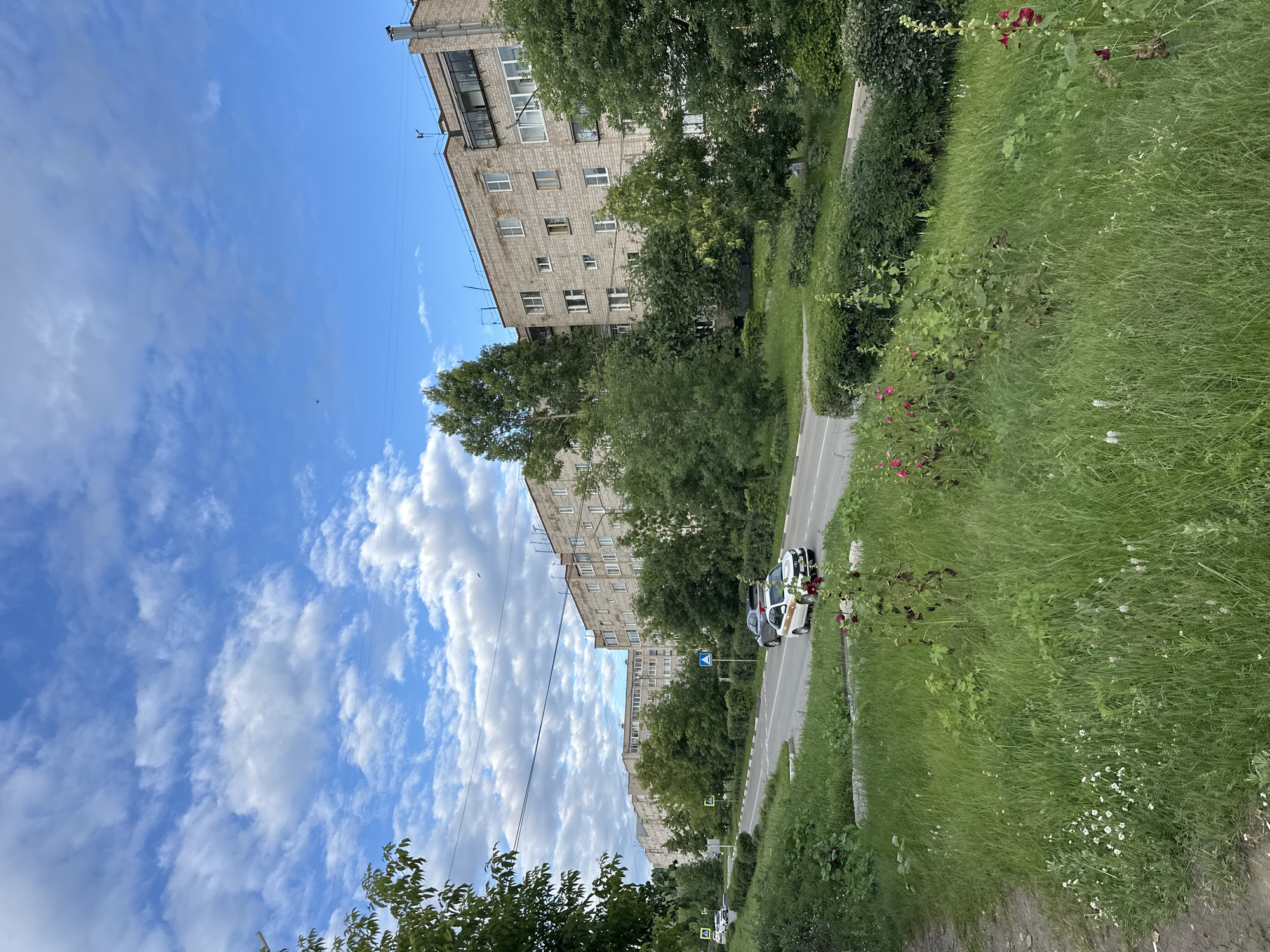 The image depicts a street scene in a residential area, likely in a city, with apartment buildings flanking a road. The buildings are made of brick and appear to be older, with multiple stories and rows of windows. Trees and lush green grass, dotted with wildflowers, line the road and the areas around the buildings, suggesting a warm season, possibly summer.
Several cars are visible on the road, and some appear to be parked. The sky is a bright blue with scattered white clouds, indicating a pleasant day. There are a few street signs visible, including a blue sign with a white symbol, and yellow pedestrian crossing signs. The perspective of the image is from a slightly elevated angle, looking down at the street and buildings.
The overall impression is one of a typical urban or suburban environment on a clear, sunny day. There are no people readily visible in the frame, and the focus is on the architecture, greenery, and infrastructure of the location. The presence of trees and grass suggests a green city or a suburban setting.