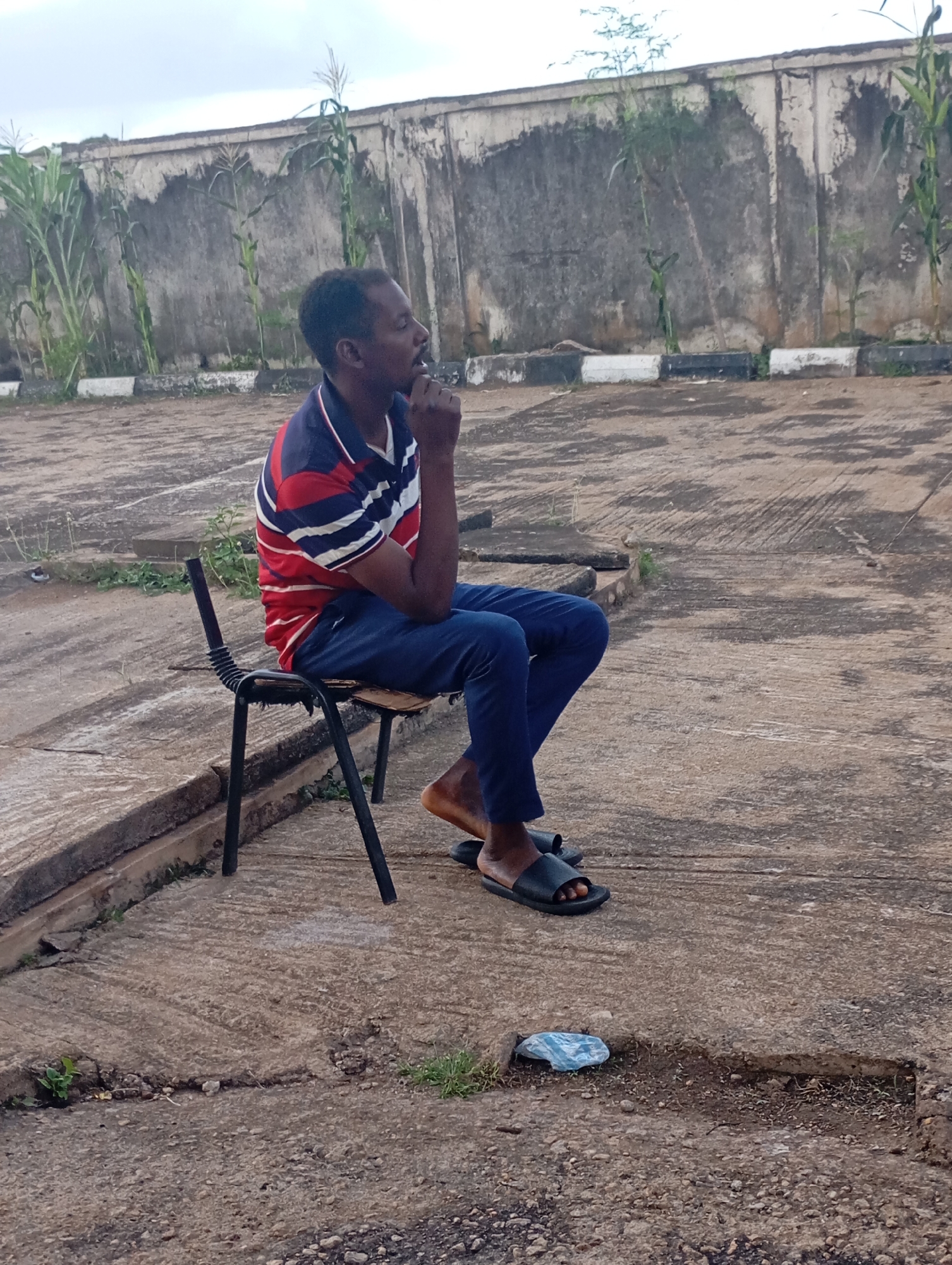 An adult Black male is seated on a black metal chair with a black plastic seat and back, facing right in profile. He is wearing a red, white, and blue striped polo shirt and dark blue pants. His left hand is resting on his chin, and he appears to be looking down or to the side. He is wearing black slide sandals. The chair is positioned on a concrete surface that shows signs of wear and has some cracks and debris. Behind the man is a rough, gray concrete wall with some vegetation, including corn stalks, growing in front of it. The wall has a deteriorated surface and some visible water damage. The ground in front of the wall has a border of alternating white and black painted concrete blocks. The sky is overcast. The scene is outdoors and appears to be in an urban or semi-urban area, consistent with a location in Shendam, Nigeria. A small piece of blue plastic is visible on the ground near the bottom right of the image.