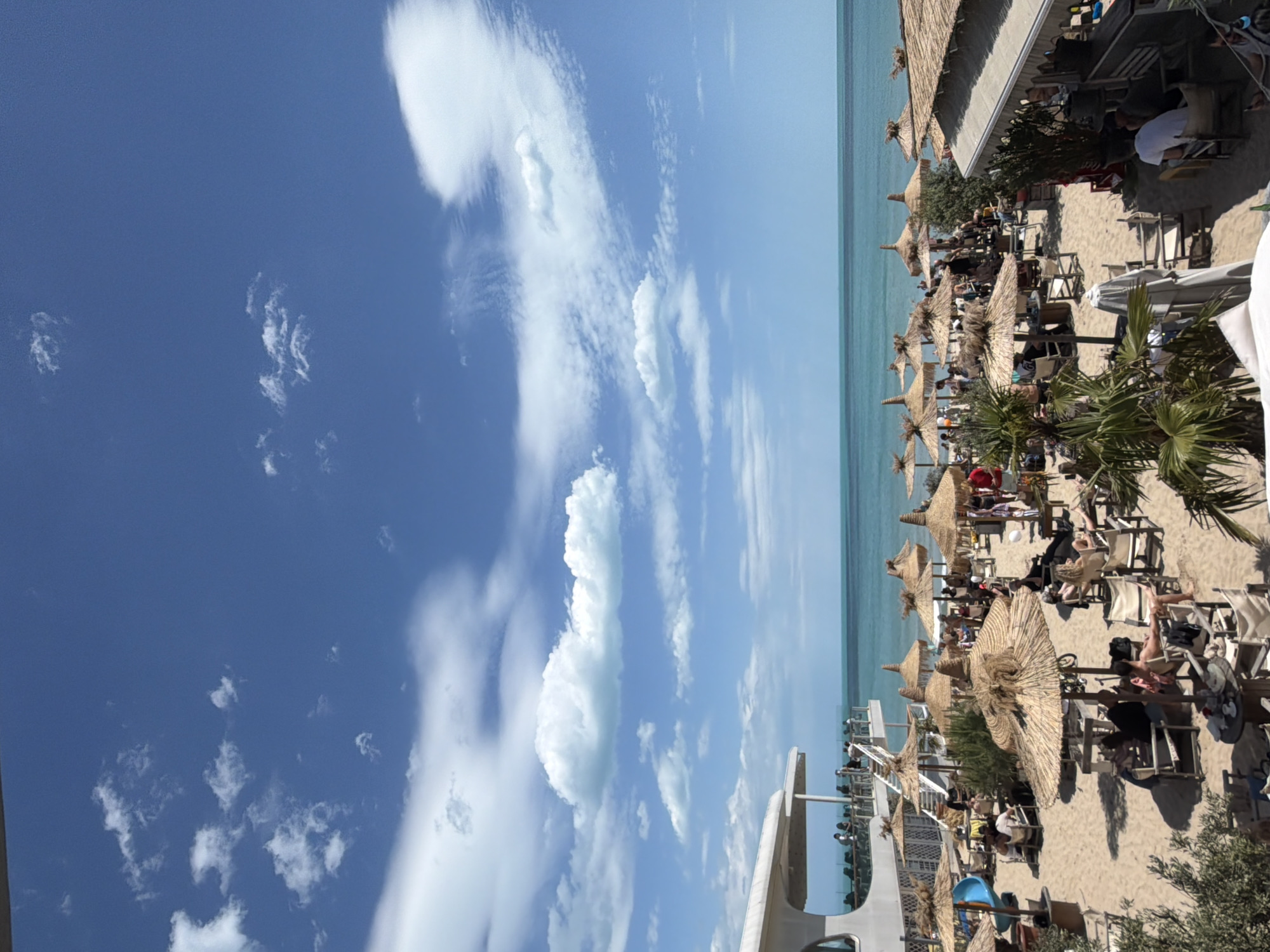 The image shows a beach scene in Varna, Bulgaria, under a bright blue sky with scattered cumulus clouds. The beach is sandy and dotted with numerous thatched umbrellas providing shade for sunbathers in lounge chairs. Some individuals are visible under the umbrellas and near the water's edge. Along the edge of the beach, there is a building, and some palm trees are near some beach umbrellas. The water appears to be calm.