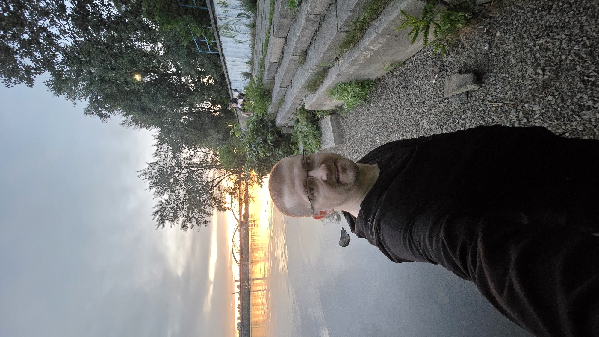 This is a selfie taken by a man with glasses, a shaved head, and a black hooded shirt. He is standing on a gravel path next to a body of water, possibly the Dnieper River in Kyiv, Ukraine. In the background, a bridge with multiple arches is visible, spanning the river at sunset. The sky is cloudy with orange and yellow hues from the setting sun reflecting on the water. To the right of the man, there are concrete steps leading up to a wall with graffiti. Two individuals are seated on the steps in the background, and trees and other vegetation are present along the riverbank and beside the steps. The overall impression is of an urban waterfront scene at dusk.