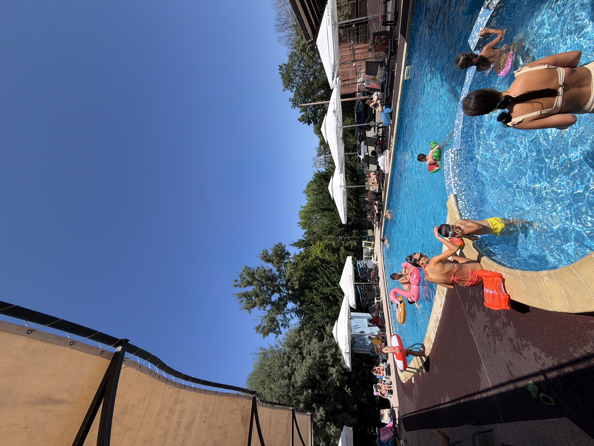 The image depicts a vibrant scene at a sunny outdoor swimming pool, likely in Burgas, Bulgaria, during the daytime. The clear blue sky suggests fair weather, perfect for a day of recreation. A group of people are enjoying the water, with several individuals swimming or floating. A child is playing with a watermelon-shaped inflatable toy, while others are relaxing on inflatable flamingos and a ring float.
Around the pool, lounge chairs are set up under white umbrellas, providing shade for sunbathers. Some people are seated on the chairs, while others are in the process of entering or exiting the pool. The pool itself is a bright blue, tiled with a mosaic pattern along the edge. The surrounding area features lush green trees and a glimpse of buildings with awnings, indicating a resort or recreational facility.
One prominent detail is the presence of orange life vests placed on the pool edge. The overall atmosphere is one of leisure and enjoyment, with people engaging in typical summer pool activities. The sunlight creates strong shadows, suggesting it is likely midday. The visible text on the umbrellas reads "ЛЕГЕНДА" which translates to "LEGEND" in Bulgarian, further confirming the location.