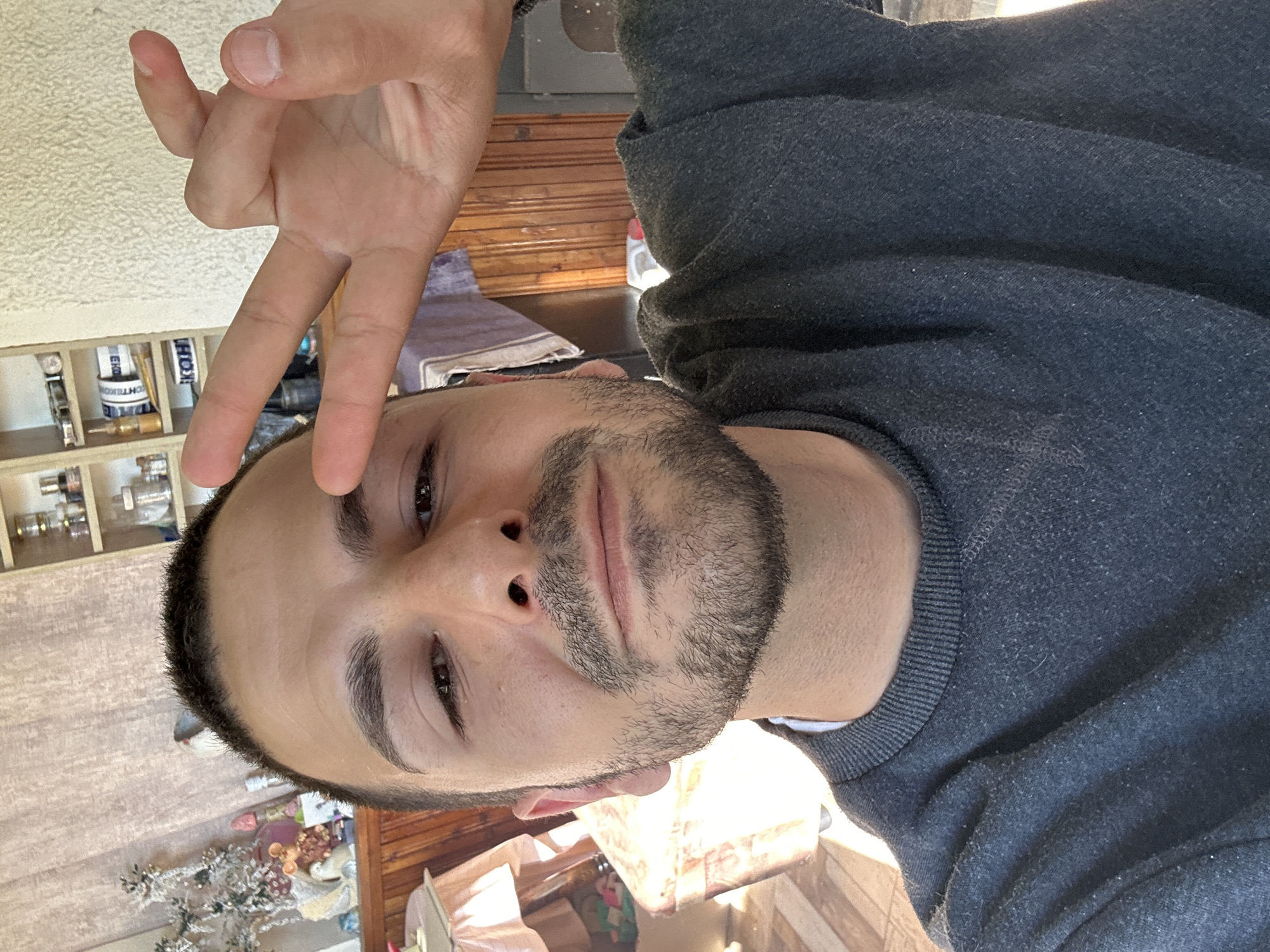 A person is making a peace sign with their hand. The person has brown hair and a beard. The person is wearing a grey sweater. There is a shelf with bottles on the left side of the image. The location appears to be indoors. The location is not in the city of Slavyani, Bulgaria.