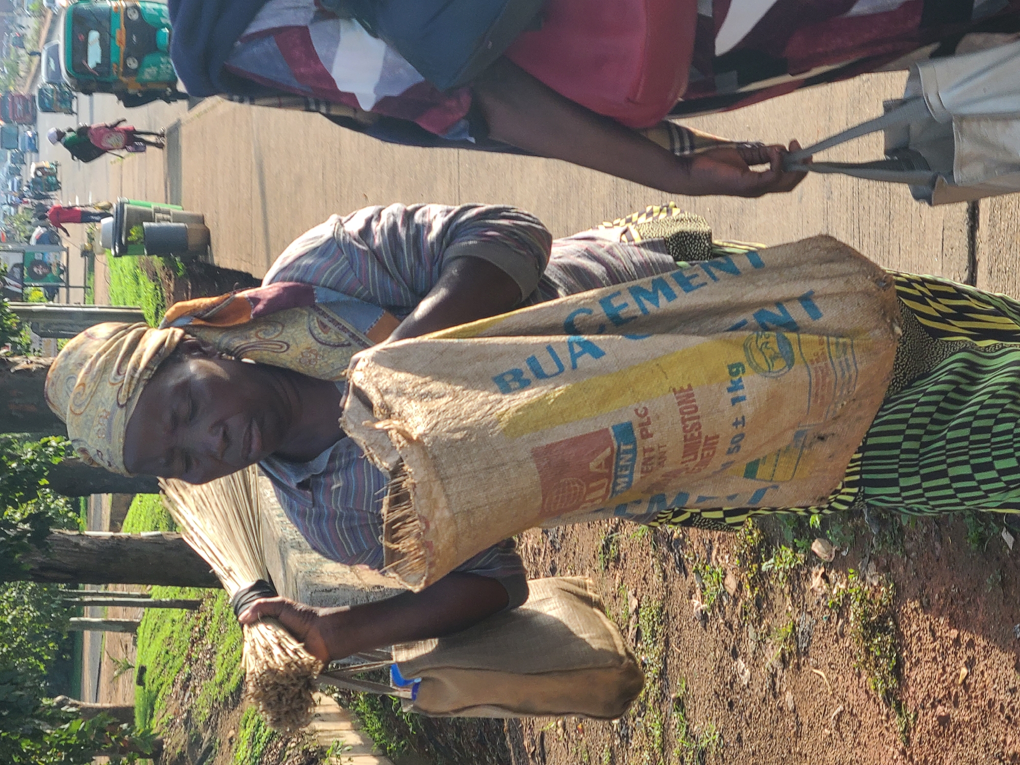 The image depicts an outdoor scene in Gura Top, Nigeria. In the foreground, a woman wearing a headscarf and a striped shirt is carrying a large burlap sack with "BUA CEMENT" printed on it, and a bundle of broomsticks. Another person, partially visible, is walking alongside her, carrying a tote bag. In the background, a busy street is visible with various vehicles, including a tuk-tuk, a truck, and cars, as well as other pedestrians. The environment appears to be a roadside with trees and vegetation lining the area. The lighting suggests it is daytime.