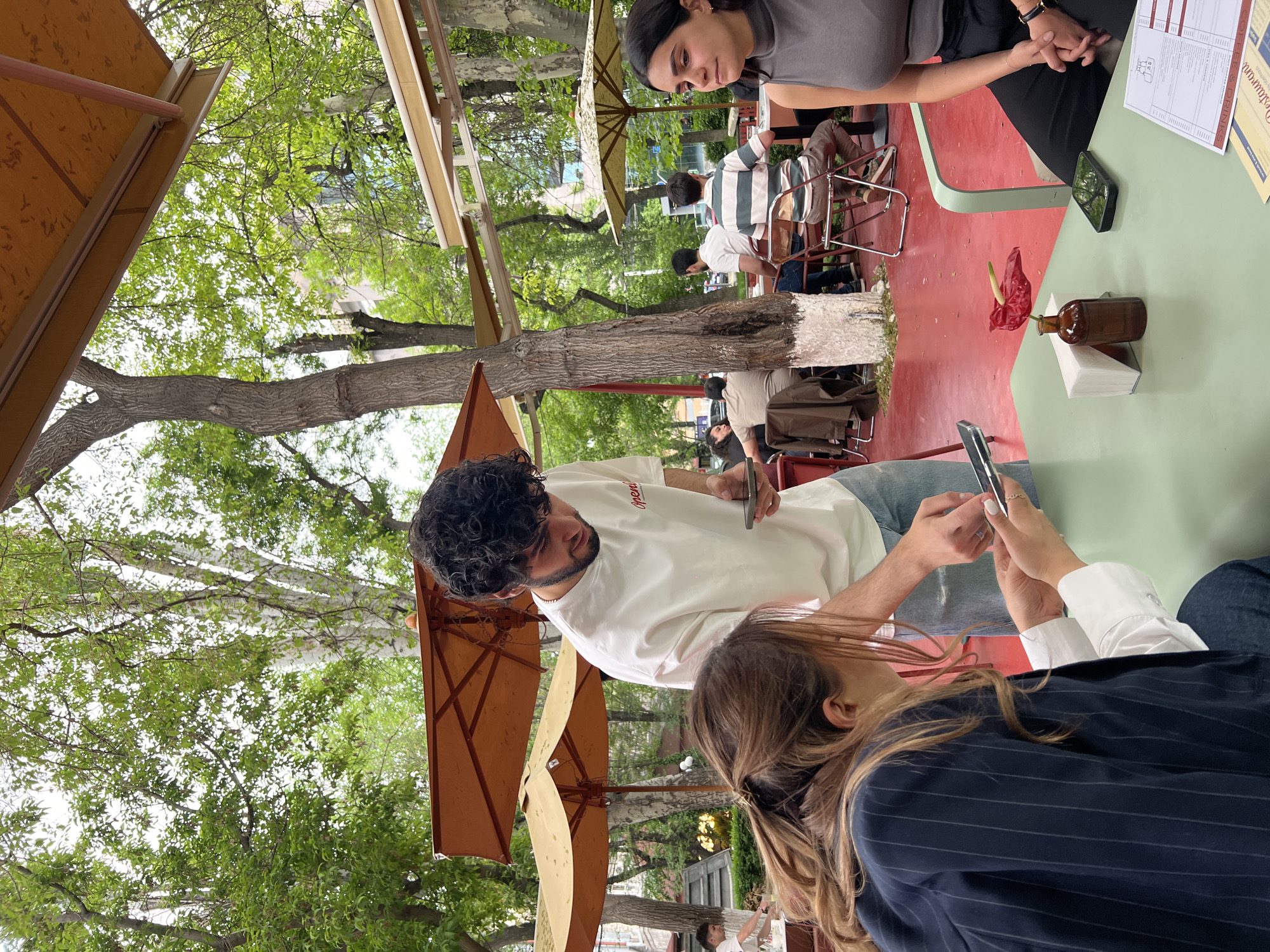 The image shows a scene at an outdoor cafe in Yerevan, Armenia. The cafe features tables with red and green surfaces, shaded by umbrellas and surrounded by trees. Four individuals are visible, each occupied with their mobile phones. A man in a white t-shirt and jeans holds a phone in one hand and a possible e-cigarette in the other. A woman in a dark blazer and jeans is seated across from him, using her phone. Another woman in a gray shirt is seated at the table. There is an arrangement of a red flower in a small bottle on the table, next to some napkins. In the background, other people are sitting at additional tables.