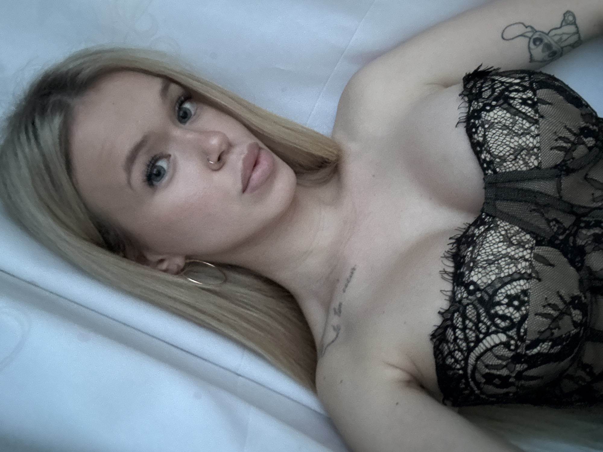 This is a medium close-up, high-angle selfie of a young Caucasian woman with blonde hair and blue eyes, wearing a black lace bra. She is lying on a white surface, possibly a bed or a couch. The woman has a nose ring and a tattoo of a cartoon character on her upper right arm. Another tattoo, possibly of text or a small design, is visible on her left shoulder. A large hoop earring is also visible in her left ear. The lighting suggests it is daytime, and the setting appears to be indoors. The woman's expression is neutral, with her eyes looking towards the camera. The focus is primarily on her face and upper body.