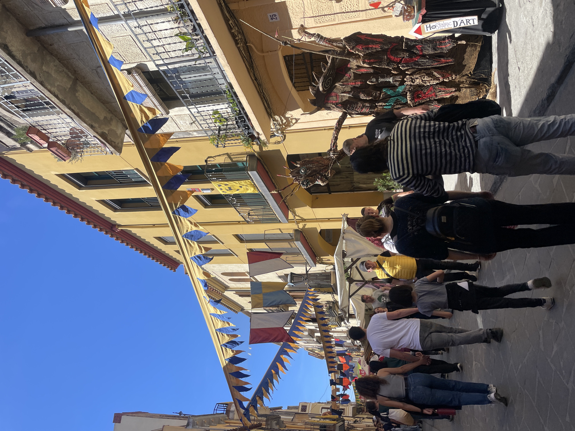 The image captures a street scene in Hostalric, Spain, likely during a festival or market day. The street is lined with yellow buildings, and pennants of various colors stretch overhead. People are walking along the street, with a vendor displaying handcrafted artwork. A sign that says "Hostalric D'Art" points to the booth.