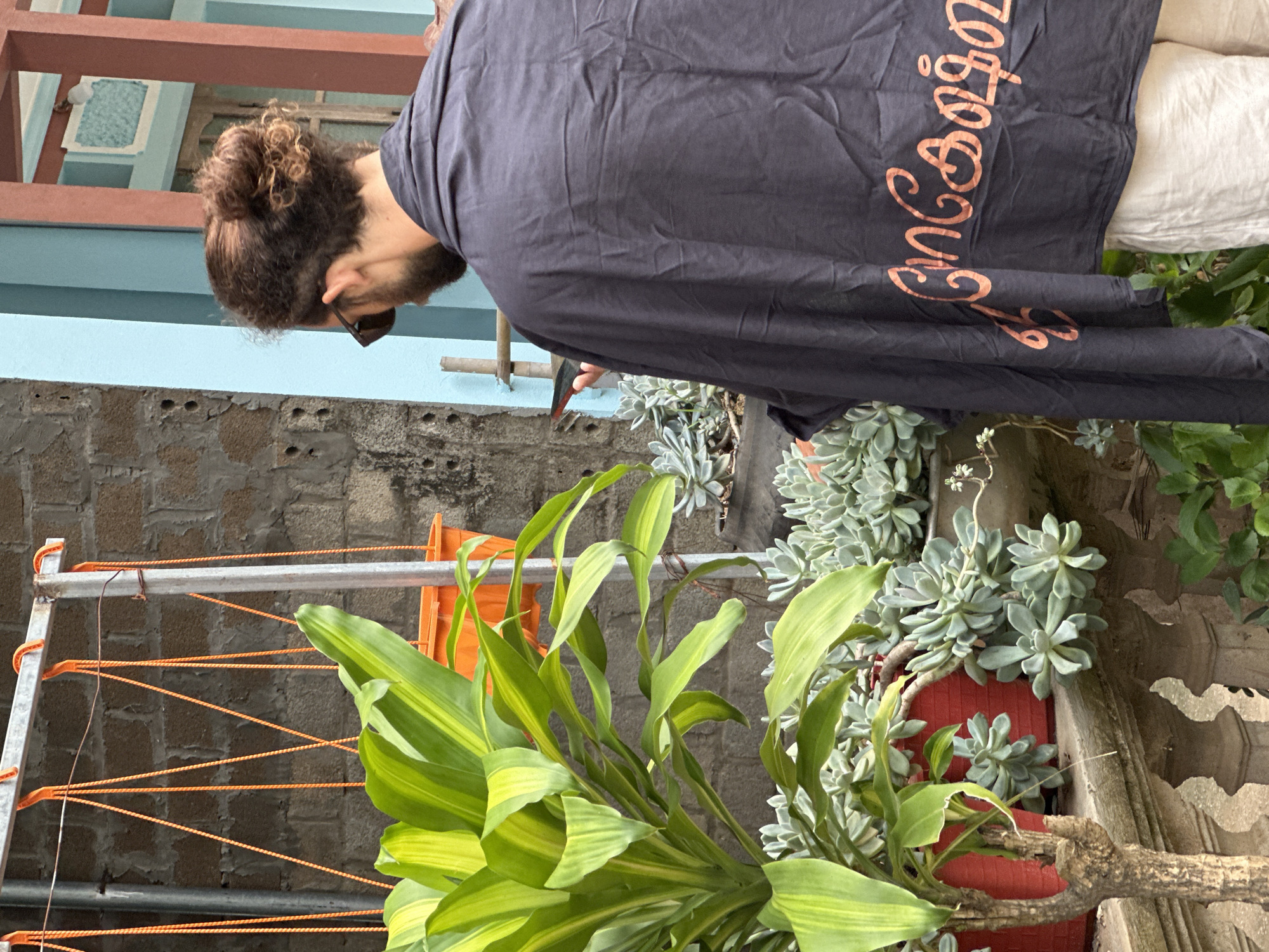 In Mai Chau, Vietnam, a person with a man bun and sunglasses stands facing away from the camera, wearing a dark grey garment with orange text on the back. They are holding a mobile phone in their left hand. The scene features several potted plants, including a tall plant with broad green and yellow leaves in a red pot, and a collection of succulents in grey pots. A rustic balcony railing and a brick wall are visible in the background, along with a clothesline with orange ropes and a metal structure. The overall environment suggests an outdoor setting in a residential area.