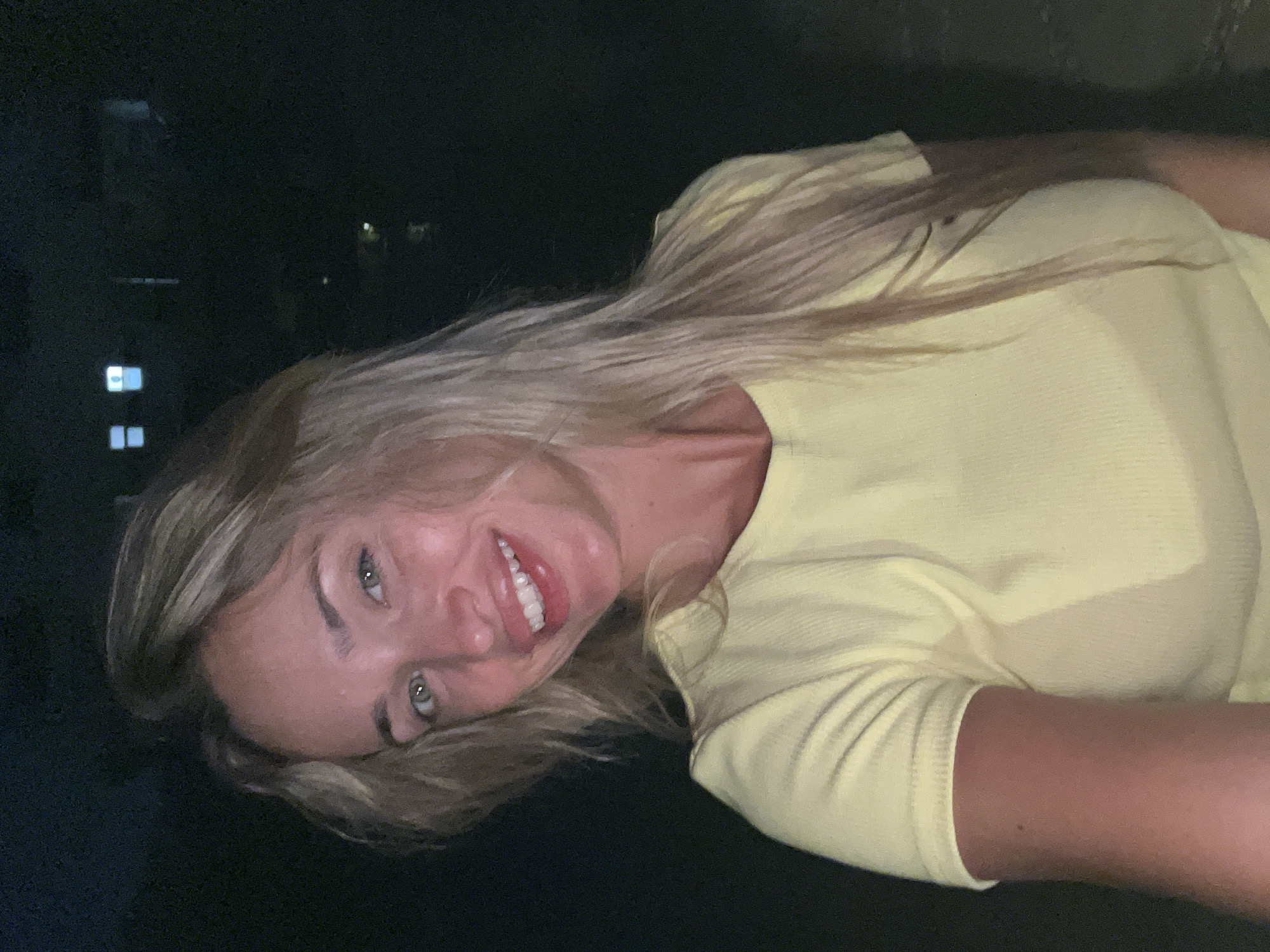 A young woman with blonde hair and light green eyes smiles at the camera, wearing a yellow t-shirt. The background is dark, with a few blurry lights suggesting buildings or a waterfront at night. It is not possible to determine the location in Sharm El Sheikh, Egypt from the provided image.