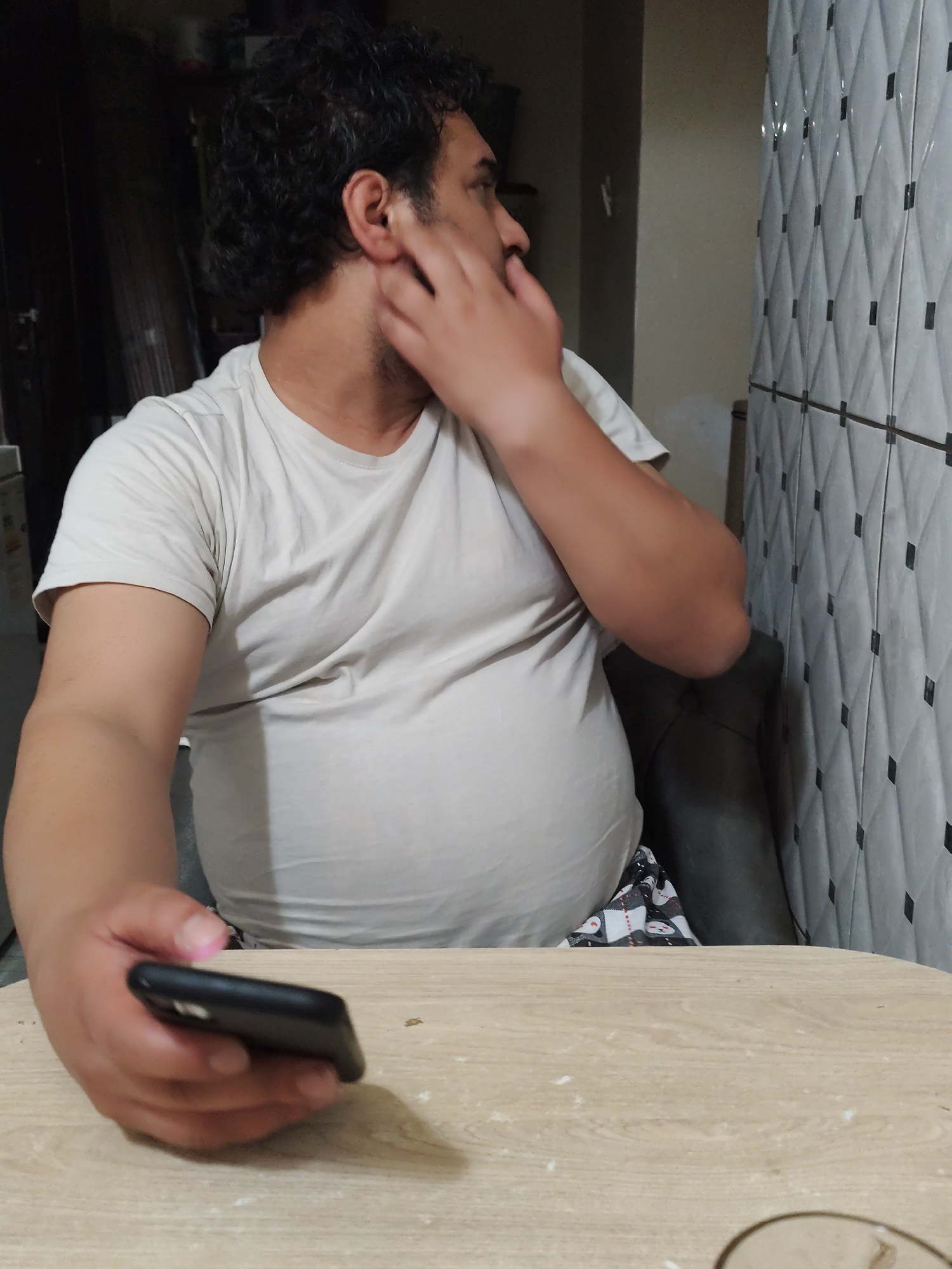 A man is sitting at a table, holding a black smartphone in his left hand. He is wearing a light-colored t-shirt. His right hand is near his face. Behind him is a wall with a diamond pattern of white tiles with black squares at the intersections. The location is Sarıyer, Turkey.