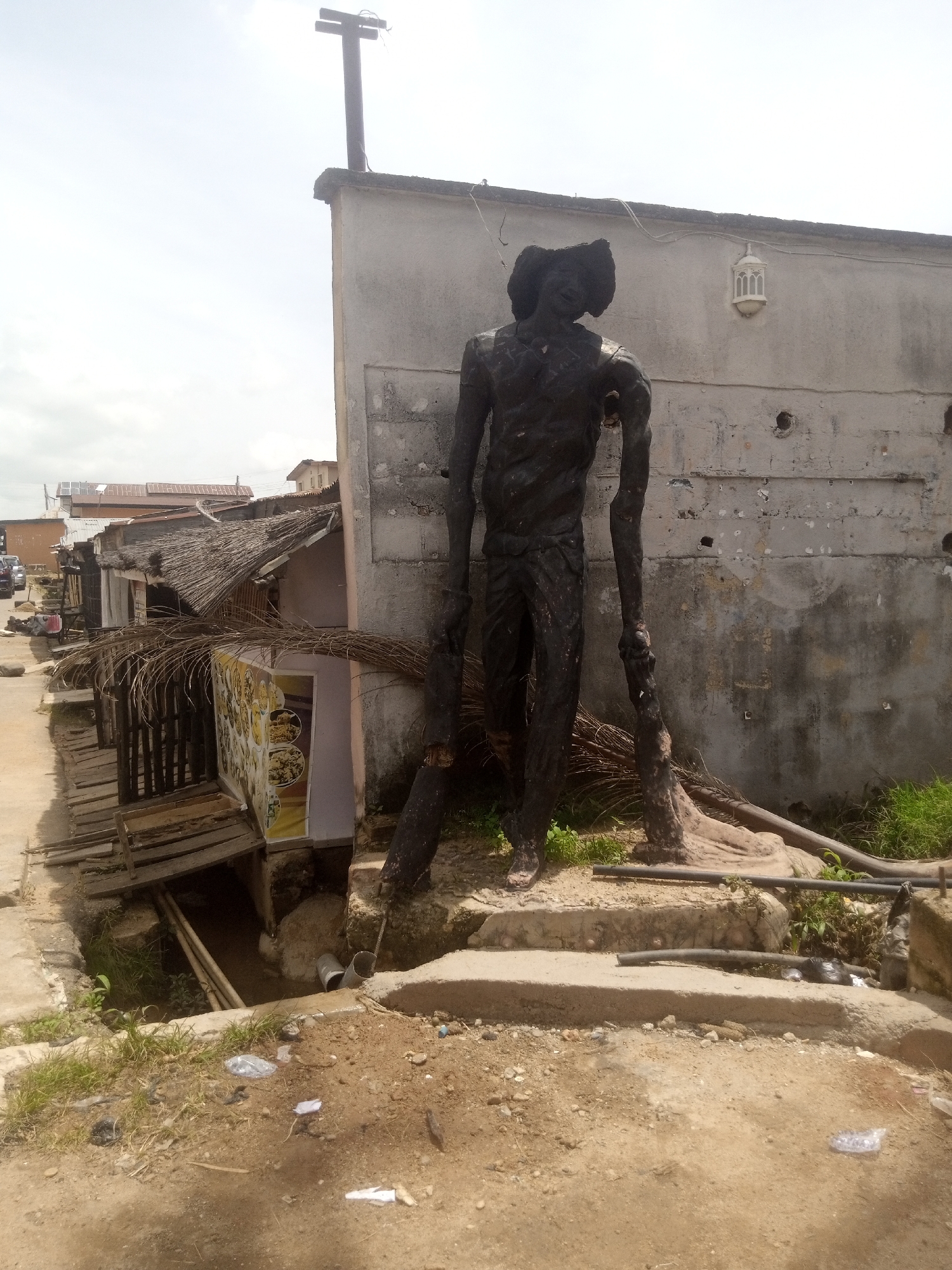 This media file showcases a statue of a human figure, rendered in a dark, rough material, standing prominently in an outdoor setting. The statue is tall and appears somewhat gaunt, with its arms outstretched, holding what seem to be large branches or logs. It is positioned on a raised, concrete platform, with a backdrop of a weathered, grey building. This building has a textured surface, with several circular holes and a decorative lantern-like fixture mounted on the wall. Atop the building, a cross is visible against the sky.
The scene appears to be a street or pathway with some dilapidated structures to the left of the statue. These structures have thatched roofs and wooden walkways, suggesting a more rudimentary environment. There's a visible drainage ditch or channel running alongside the pathway, with pipes and debris present. Some greenery, including grass and dried palm fronds, is scattered around the statue and the surrounding area, indicating an outdoor environment.
The overall impression is one of an urban or semi-urban setting that shows signs of wear and perhaps neglect. The lighting suggests it is daytime, with an overcast or cloudy sky. There is no clear indication of specific activity or event taking place, other than the static presence of the statue and the general ambiance of the location. The statue itself, with its stark form and pose, might evoke a sense of stoicism or resilience. There is a sign visible on one of the structures to the left, featuring images of food, but no discernible text can be read clearly.