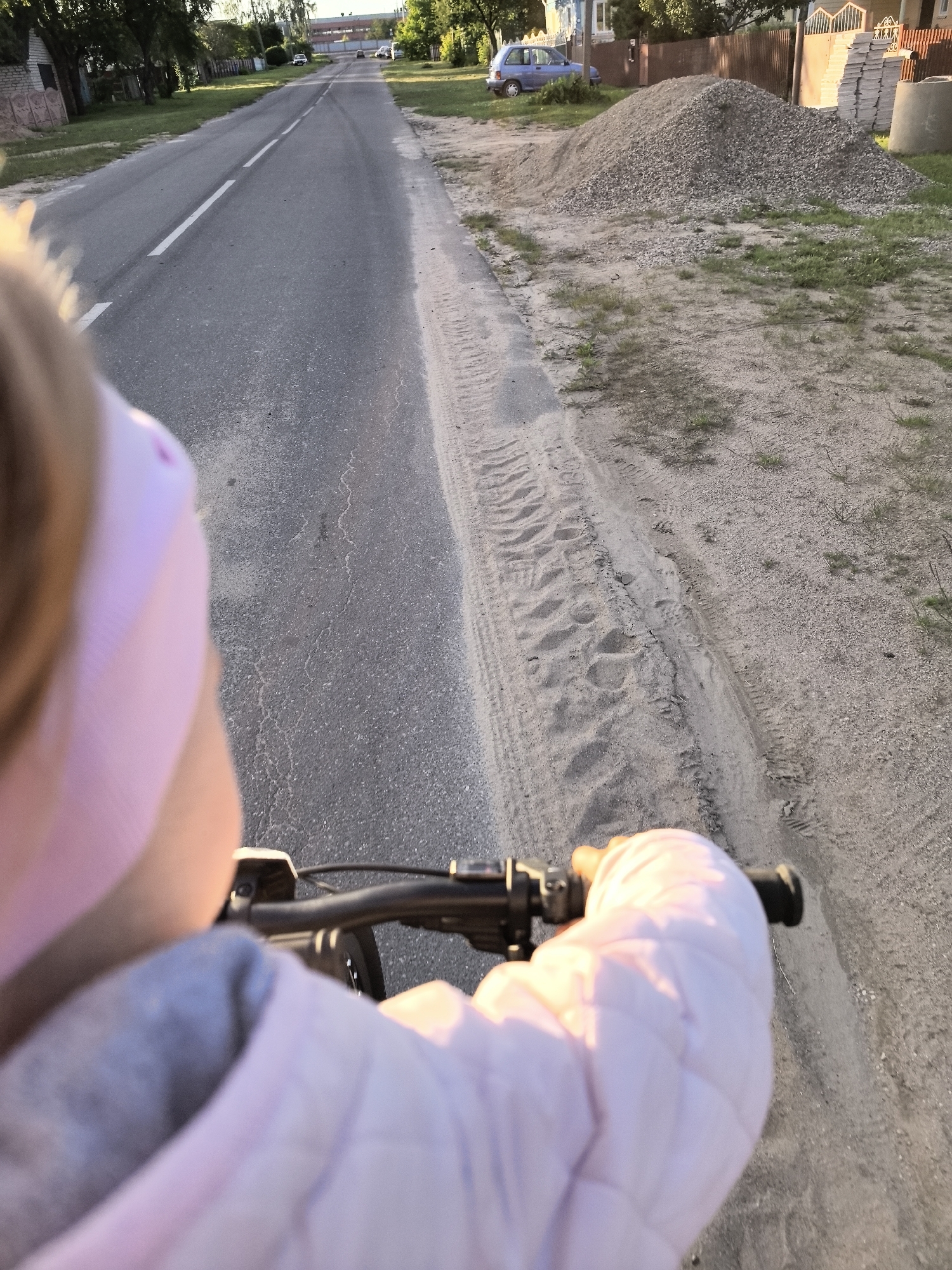 A close-up, over-the-shoulder view of a person, likely a child, wearing a pink headband and a pink quilted jacket, riding a bicycle on an asphalt road. The road has a white dividing line and tire tracks on the adjacent sandy shoulder. In the background, there are trees, houses, a pile of gravel, stacked building materials, and a concrete pipe. A blue car is parked on the left side of the road. The lighting suggests it is late afternoon.