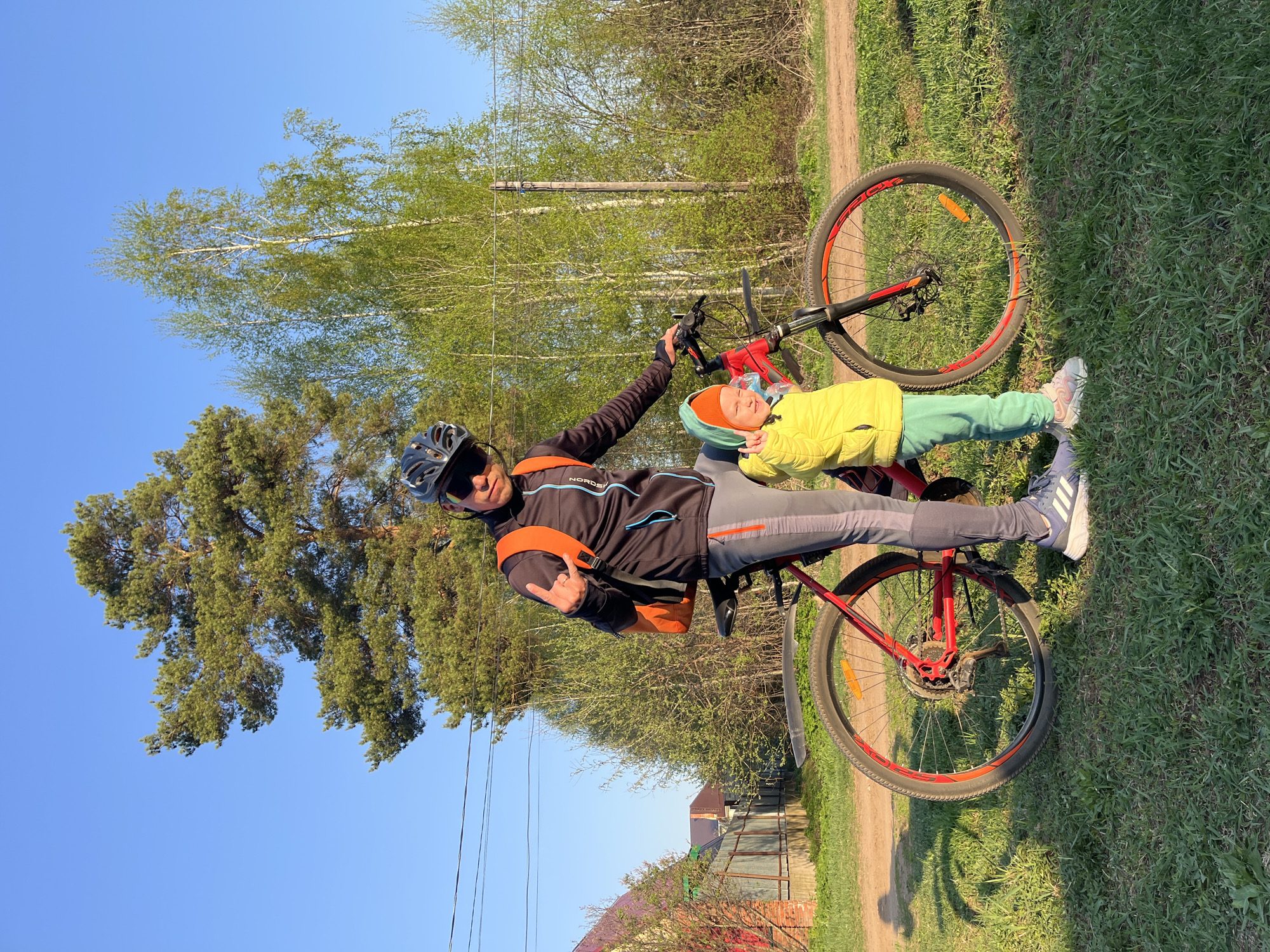 A man and a child lie on a red bicycle in an outdoor setting. The man, wearing a black cycling jacket, gray pants, a black helmet, sunglasses, and an orange backpack, extends his arm towards the camera, making a gesture with his fingers. The child, wearing a yellow jacket, green pants, an orange hat, and green head covering, is lying next to the man on the bicycle's frame. The bicycle rests on green grass, and a dirt path is visible in the background. Trees and a blue sky frame the top left corner of the image, while a fence and building are visible towards the bottom right. Based on the landscape and provided location information, this scene is likely in the Chaikovsky, Russia area.