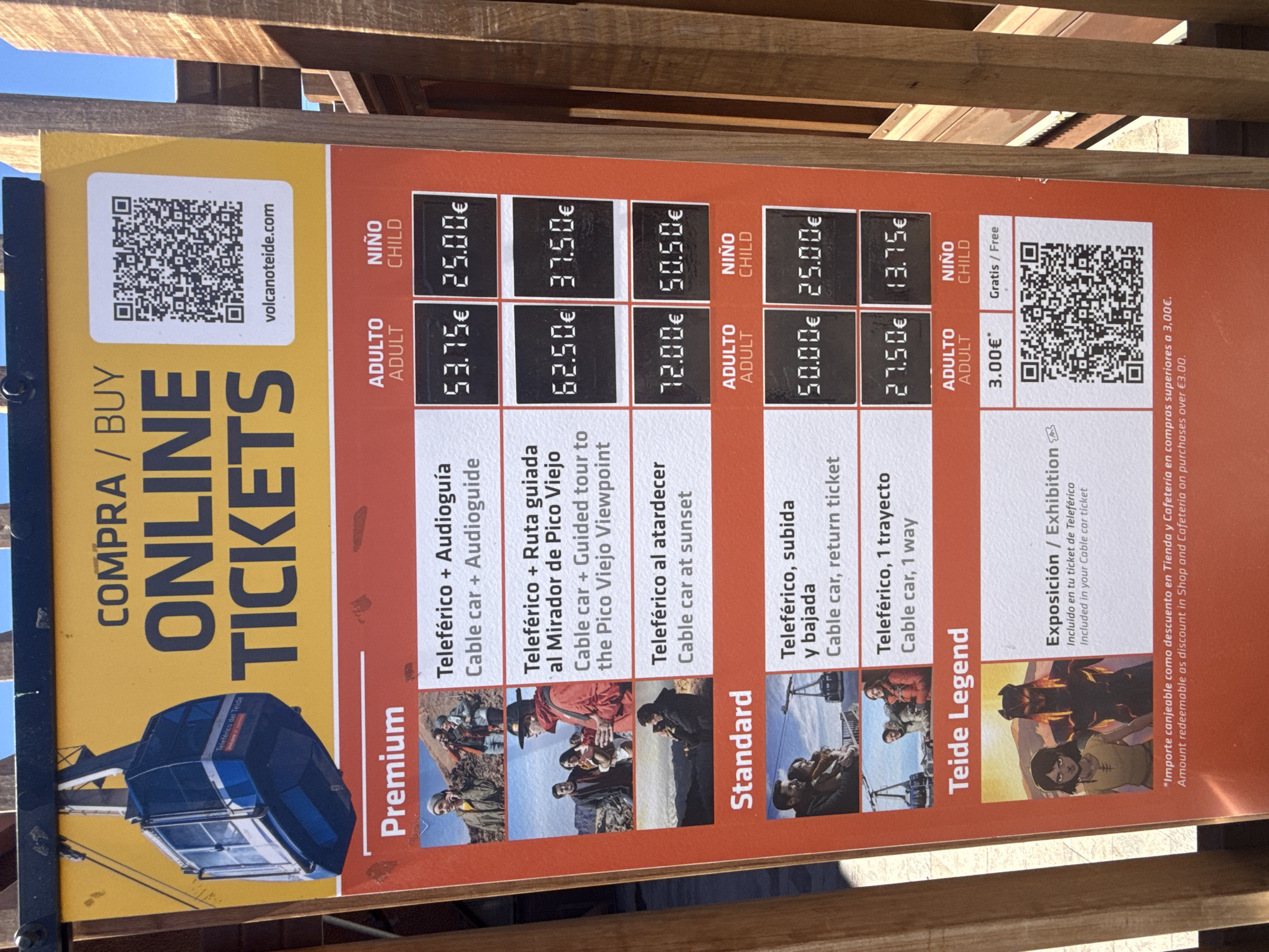 The image shows a colorful price list for the Teide Cable Car in La Orotava, Spain. The sign is predominantly yellow and orange, featuring images of the cable car, people, and a stylized drawing. It lists various ticket options categorized as "Premium" and "Standard," with prices for adults and children. A QR code is prominently displayed for online ticket purchases. The overall presentation is vibrant and informative, aimed at attracting tourists to the attraction.