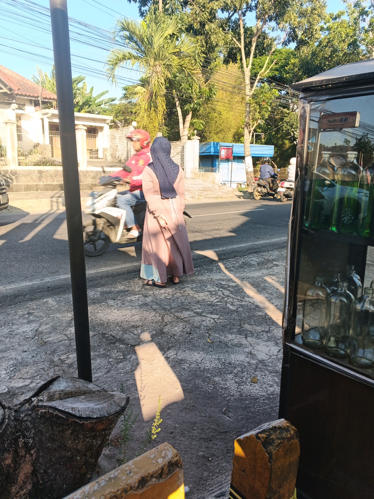 This image depicts a street scene in Semarang, Indonesia, bathed in the warm light of the late afternoon sun. The perspective is from a sidewalk, looking across a paved road.
In the foreground, on the left, a dark, weathered tree stump is visible, casting a distinct shadow. To the right, a dark display case with glass doors stands, showcasing rows of glass bottles, some filled with a green liquid.
Across the road, a woman in a long, peach-colored dress and a dark blue hijab stands facing away from the camera. Behind her, a person on a motorcycle, wearing a red jacket and a helmet, is moving away from the viewer. Further down the road, another person on a motorcycle is visible, this one in a blue shirt and helmet.
The environment includes a residential building with a tiled roof and white walls to the left of the road. Lush green trees and palm trees line the background, indicating a tropical or subtropical climate. Overhead, a tangle of electrical wires is visible against the clear blue sky. The road surface appears to be asphalt, with some cracks and debris. The overall impression is one of a typical urban street in a warm, sunny climate.