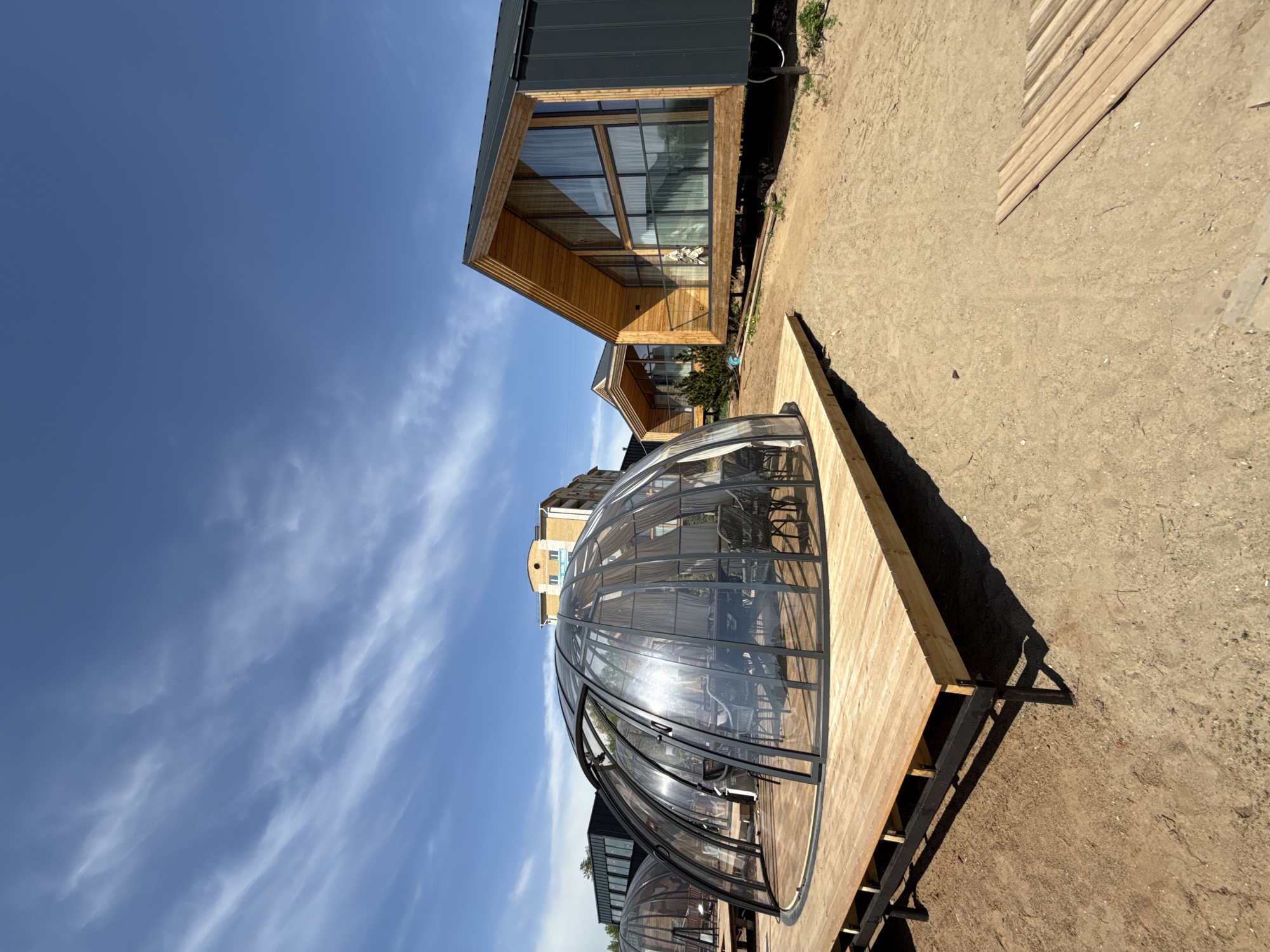 The image shows a scene in Votkinsk, Russia, featuring a cluster of modern architectural structures under a bright sky with wispy clouds. A spherical, glass-domed structure on a wooden platform is prominent, appearing to be a partially transparent, potentially habitable or dining space. Adjacent to it are contemporary wooden buildings with large windows. The ground is a mix of dirt and sandy soil, suggesting an outdoor or landscaped environment. The scene appears to be in daylight with the sun reflecting off the glass dome. The architecture combines modern design with natural materials.