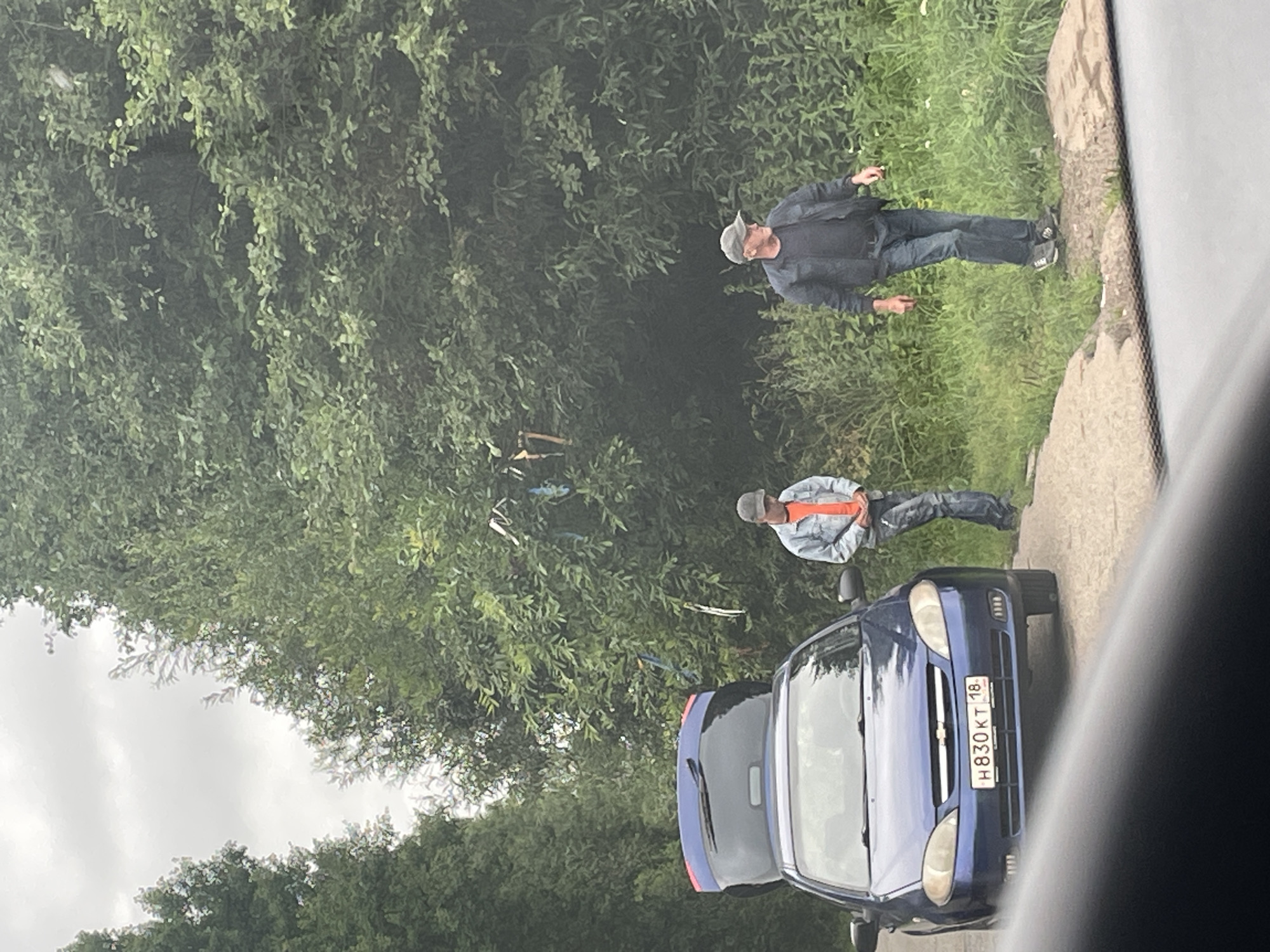 In Gavrilovka, Russia, a blue Chevrolet sedan with its trunk open is parked on the side of a dirt road. Two men are standing near the vehicle. The man on the left, wearing a light blue denim jacket over an orange shirt and blue jeans, stands by the car with his hands clasped in front of him. The man on the right, dressed in a dark jacket, blue jeans, and a grey baseball cap, stands a few feet away, looking to his left. They are surrounded by lush green foliage, with trees and tall grass forming the backdrop. The sky is overcast, suggesting a cloudy day. A cluster of colorful ribbons are tied to the branches of a nearby tree. The license plate of the car is visible and reads "H830KT 18".
