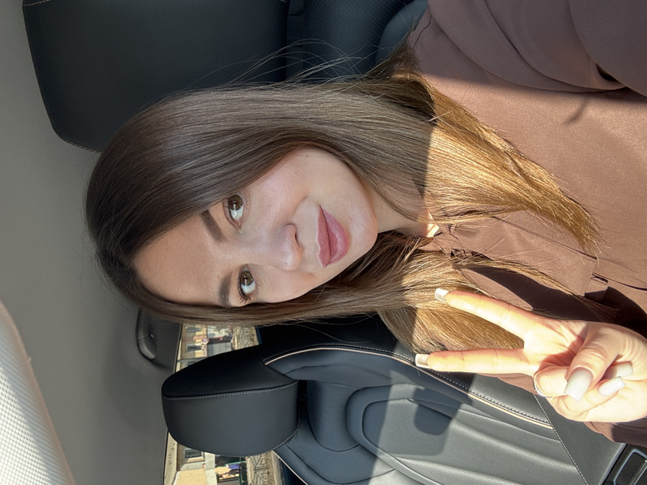 A female subject is depicted in a close-up, chest-up shot taken inside a vehicle, identified as being in Likino-Dulyovo, Russia. The subject, appearing to be in her 20s or 30s, has medium-length brown hair, brown eyes, and a light complexion, with light pink lipstick on her lips. She is wearing a brown long-sleeved top. Her right hand is raised, making a "peace" sign (V-sign) with the index and middle fingers extended. Her fingernails are square-shaped with light-colored polish, and a silver ring is visible on her right ring finger. She maintains direct eye contact with the camera and exhibits a faint, closed-mouth smile. The interior of the vehicle features black leather seating with light-colored stitching, visible on the headrest and seat to her left. Sunlight illuminates the scene, casting shadows across her hair and right arm. Through the vehicle's window, behind the car's headrest, several light-colored, multi-story residential buildings are visible, one of which appears unfinished or in a state of disrepair. The ground outside appears unpaved.