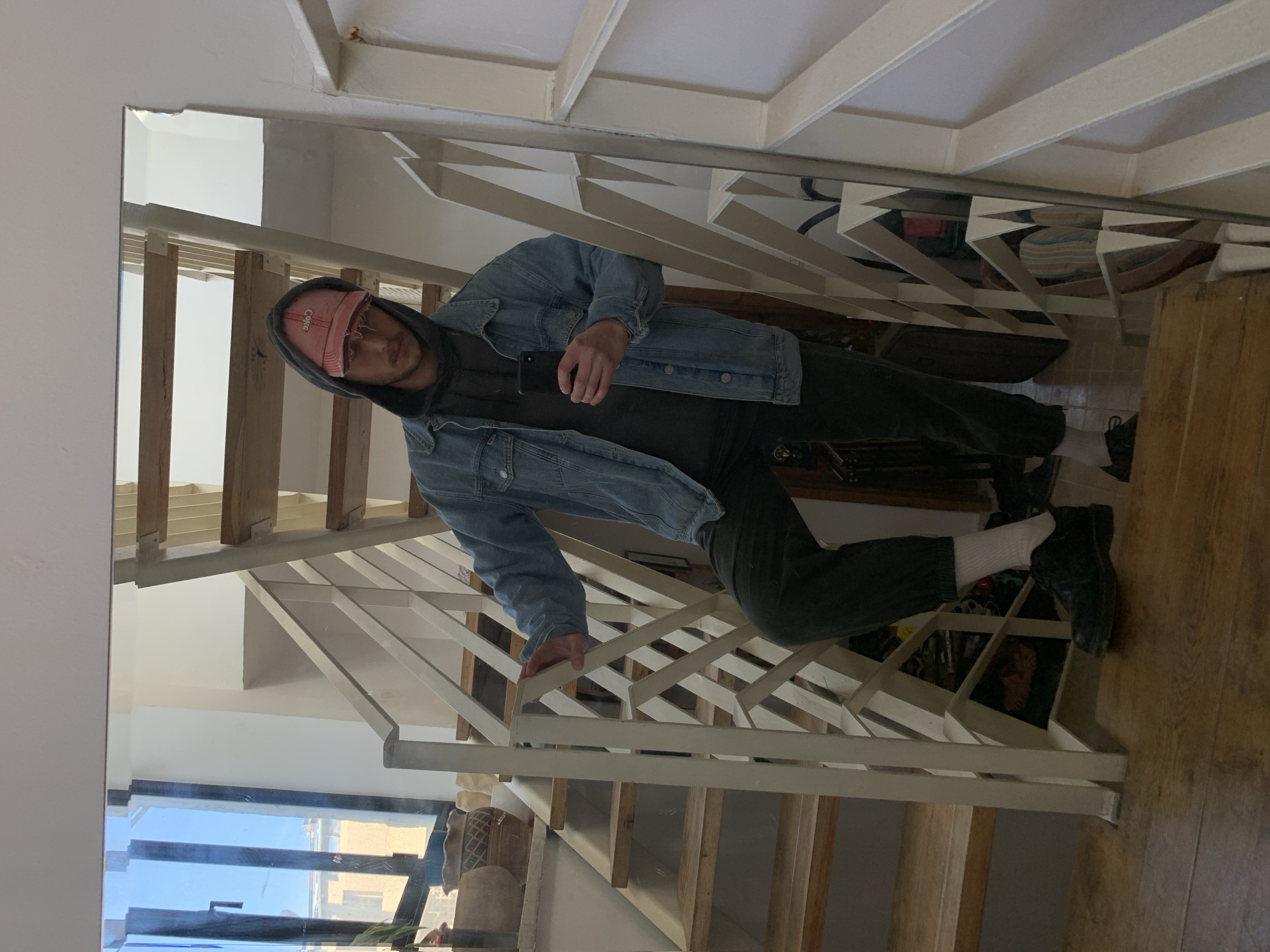 A male individual is depicted in a full-length mirror reflection, positioned on a wooden staircase within an indoor residential setting, consistent with an urban environment in Tel-Aviv, Israel.
The subject is standing with his left foot on a lower step and his right foot on a step above, suggesting a pause during ascent or descent. His left hand is resting on a white metal banister with a geometric, criss-cross design. In his right hand, he holds a black smartphone, oriented vertically, indicating he is taking the mirror selfie.
He is dressed in a dark gray hooded sweatshirt, with the hood pulled up, and a light blue denim jacket worn over it. He wears dark green or black jogger-style pants, white crew socks, and black lace-up shoes. A distressed pink baseball cap, featuring the word "Coke" in reversed red lettering, is worn over the hood. His facial expression is neutral, and his gaze is directed towards the camera/mirror.
The staircase features light-toned wooden steps. Behind the subject, a large window with dark frames reveals an exterior view consisting of a clear blue sky and light-colored building structures with vertical elements. On a windowsill or shelf beneath the window, several decorative pots or vases are visible, one being a textured terra cotta brown and another a darker, patterned vessel, accompanied by green foliage. The walls of the interior are painted white.