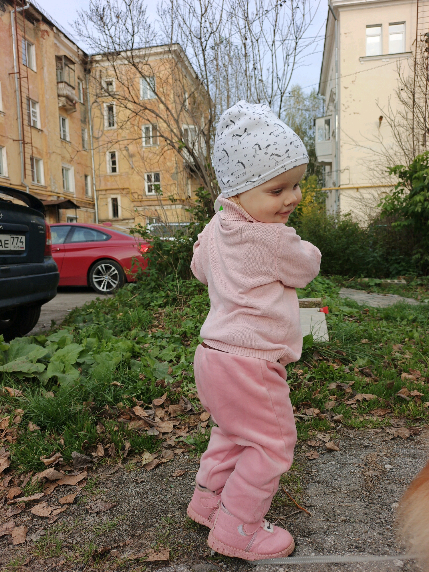 A young child, wearing a pink sweater, pink pants, pink boots, and a white beanie with a unicorn pattern, stands on a gravel path surrounded by grass and fallen leaves. The child is facing away from the camera, looking to the right. In the background, a red car is parked next to a dark-colored vehicle. Behind the cars, two apartment buildings are visible, one appearing older and painted yellow with a rusted fire escape, and the other newer and cream-colored. The setting appears to be a residential area in Zlatoust, Russia, likely a courtyard given the presence of parked cars and apartment buildings. The sky is overcast.