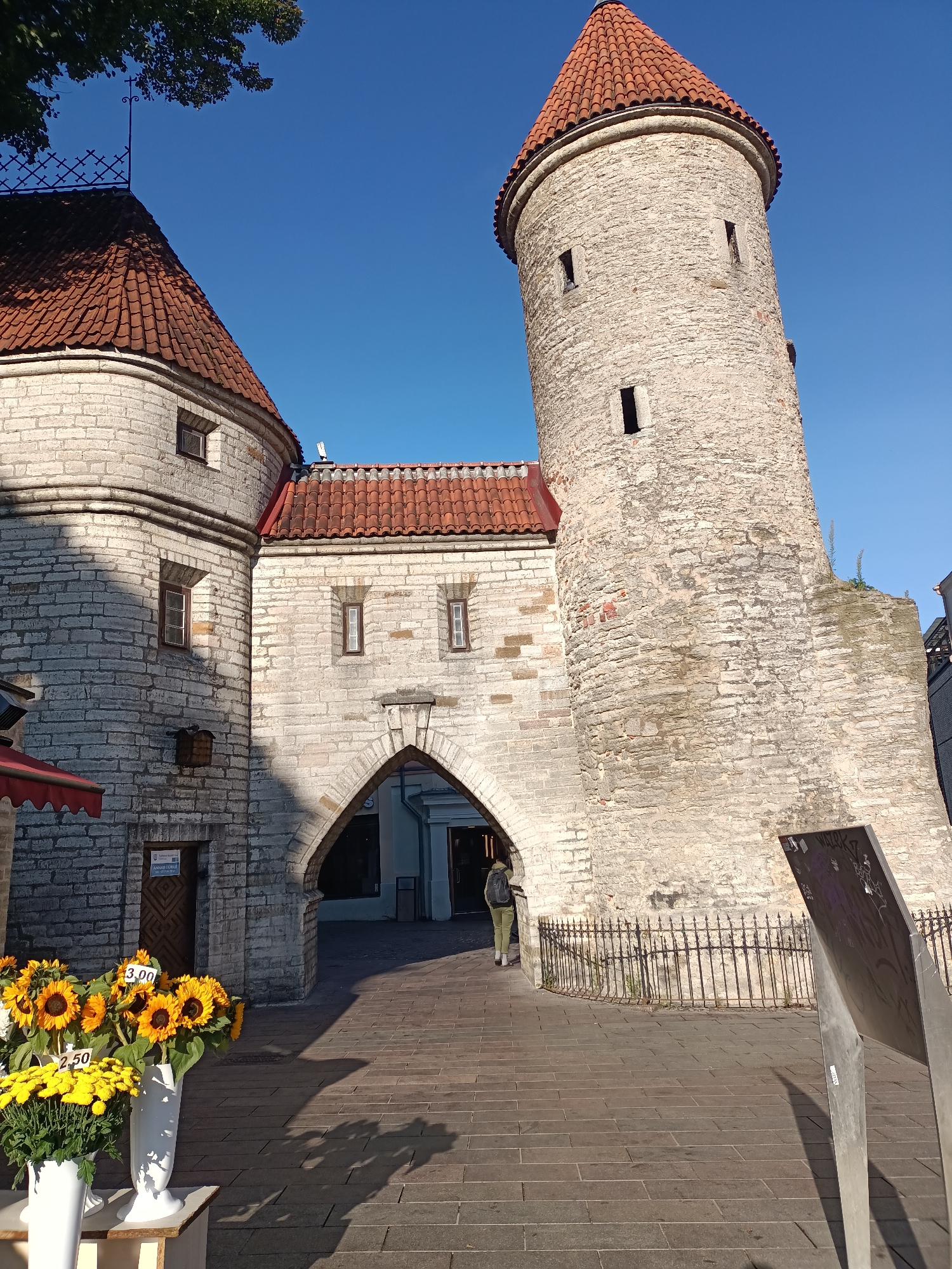 The image shows a view of the Viru Gate in Tallinn, Estonia. The gate is a historic stone structure with two round towers topped with red conical roofs. An arched passageway leads through the gate. In the foreground, on the left, are two vases of sunflowers and yellow chrysanthemums for sale, with price tags visible. A person wearing a green jacket and backpack is walking through the arched passage. The ground is paved with stone. The sky is clear and blue. The overall scene is bright and sunny.