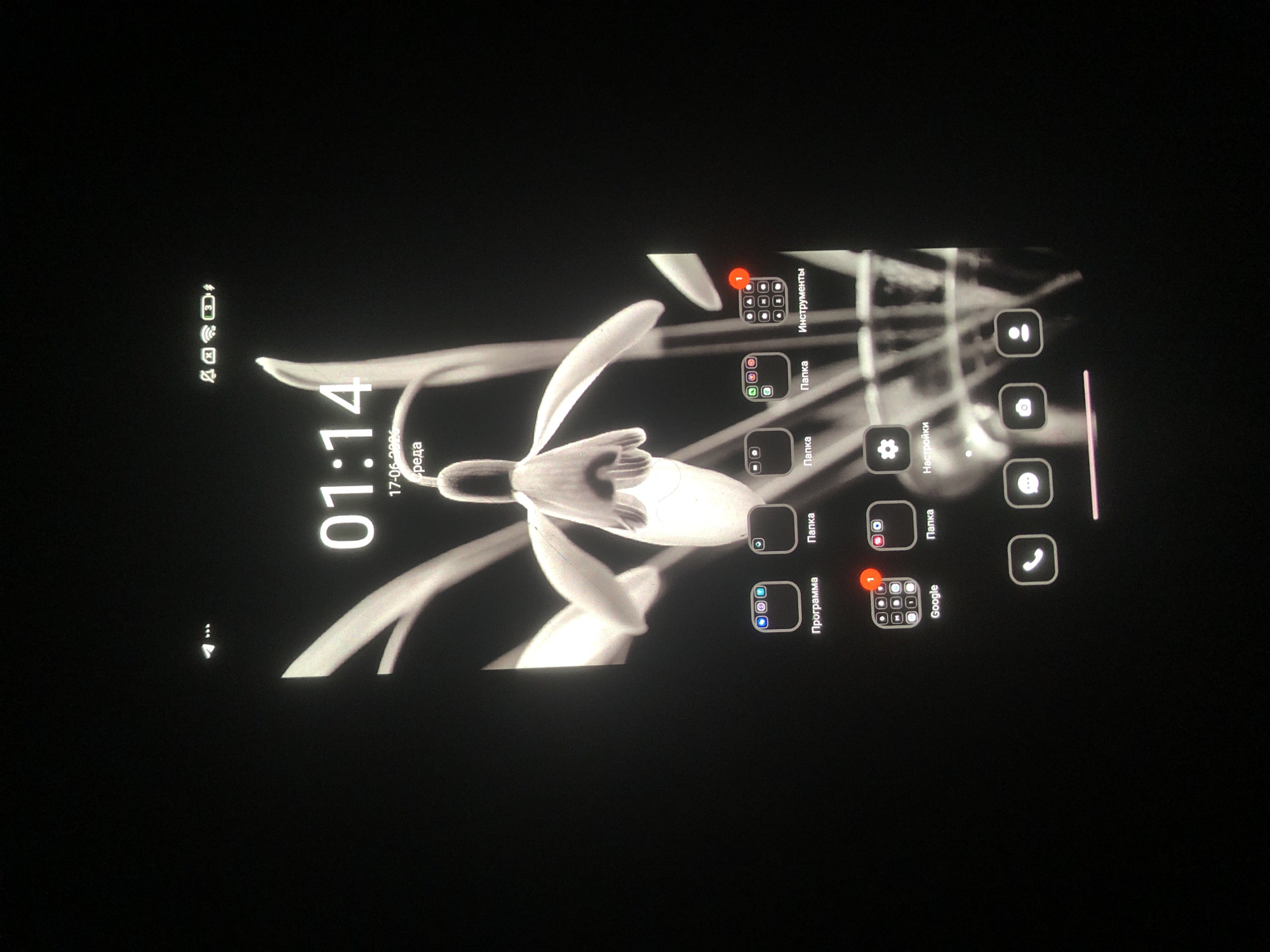 This is a close-up, black and white image of a smartphone screen displaying a lock screen. The background features a stylized, glowing image of a snowdrop flower. Prominently displayed in the center is the time "01:14" along with the date "17-06-2021" and the day of the week "среда" (Wednesday in Russian). Below the time, there are several app icons arranged in rows. The top row includes icons labeled "Программа" (Program), "Папка" (Folder), and "Папка" (Folder), with another icon labeled "Инструменты" (Tools) partially visible on the right. The second row shows a "Google" icon with a notification badge, a "Папка" (Folder) icon, and a "Настройки" (Settings) icon. The bottom row contains icons for phone, camera, and contacts. The status bar at the top shows icons for signal strength, Wi-Fi, battery level, and other notifications. The image does not provide any specific visual cues to determine its location within the city of Tashkent, Uzbekistan.