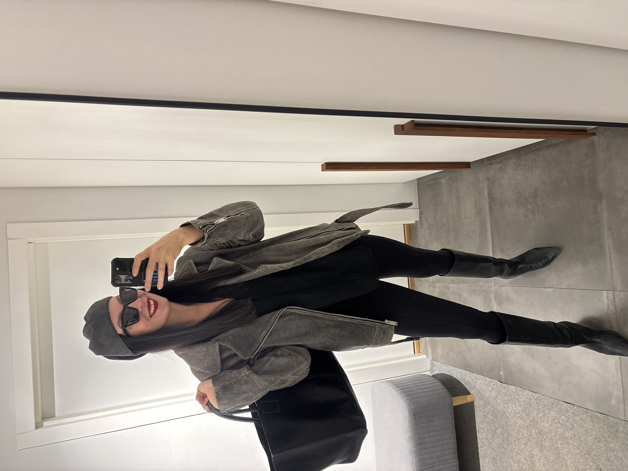 A woman is taking a selfie in a well-lit interior. She is wearing a gray beret, sunglasses, a gray jacket, black top, black pants, and black knee-high boots. She is holding a black smartphone in her right hand and a black tote bag in her left. The background shows a white wall, gray tiled floor, and a gray ottoman. The environment appears to be a fitting room or a showroom of some kind, with visible wooden wall fixtures near the ceiling. There is no information available to indicate this image was taken in Chaikovsky, Russia.